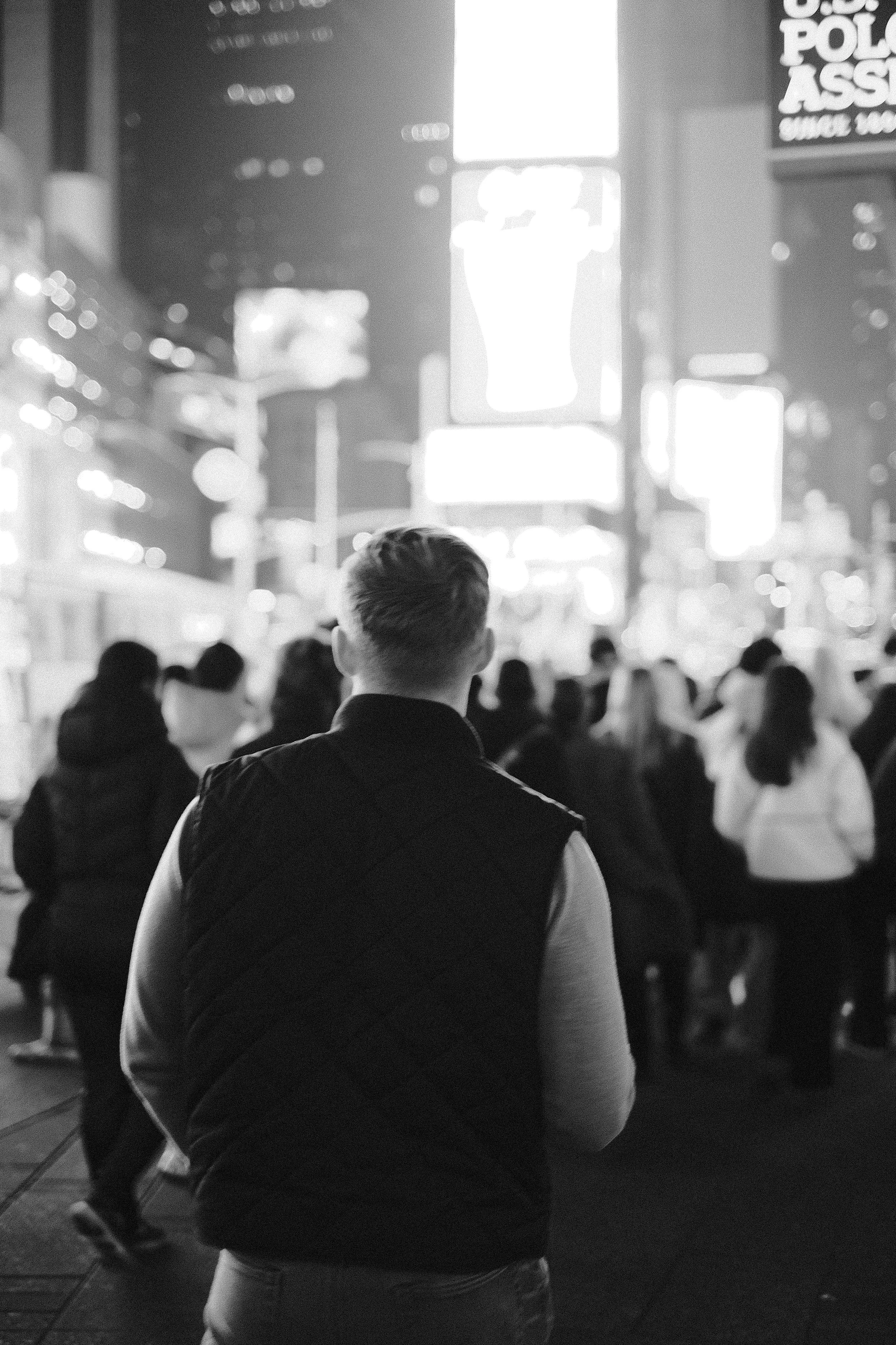 A person with short hair wearing a puffy vest standing in a busy city street at night, illuminated by bright digital billboards and neon lights, with other pedestrians around.