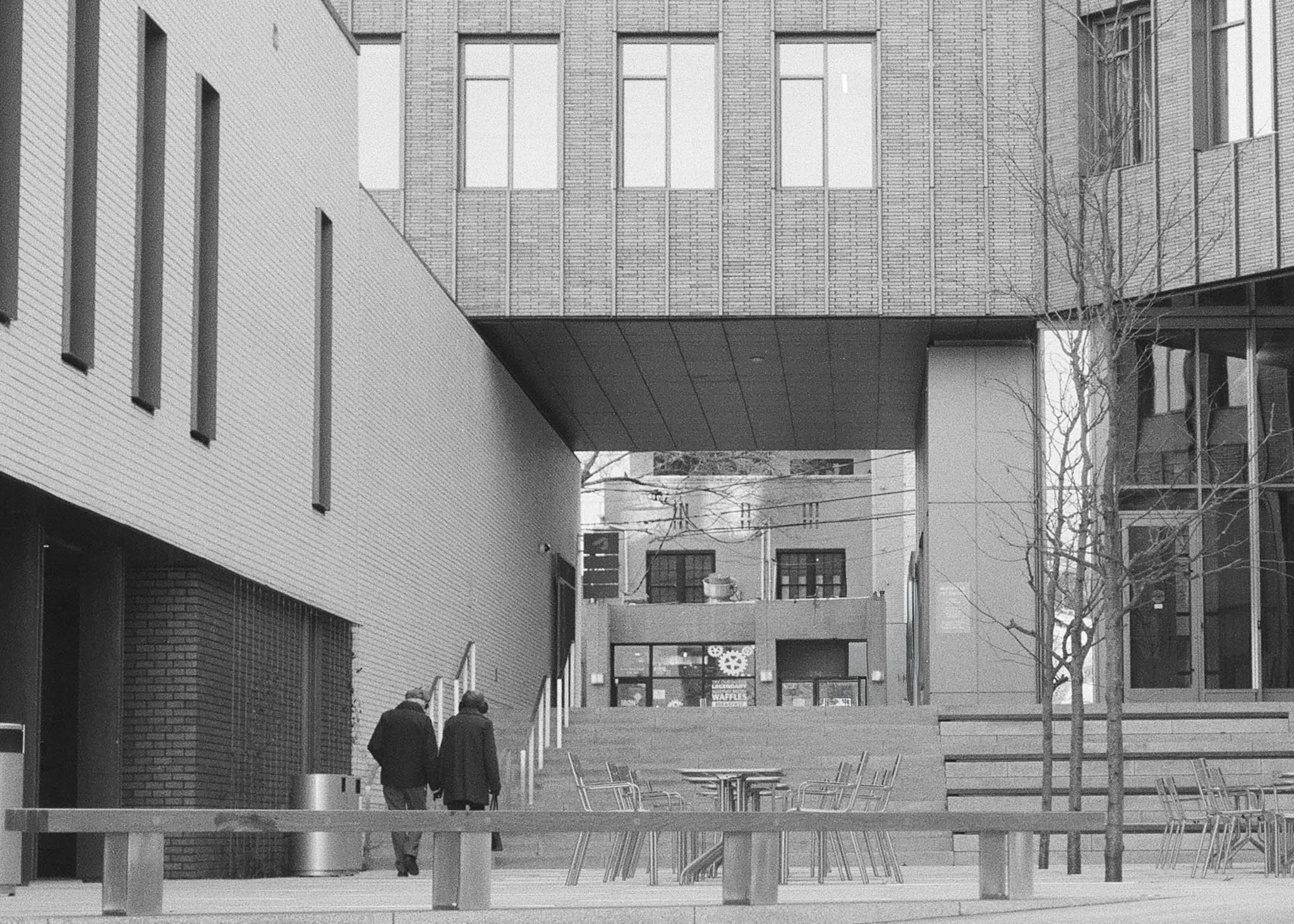 Black and white photo of two people walking in an urban area with modern buildings, stairs, benches, and leafless trees.