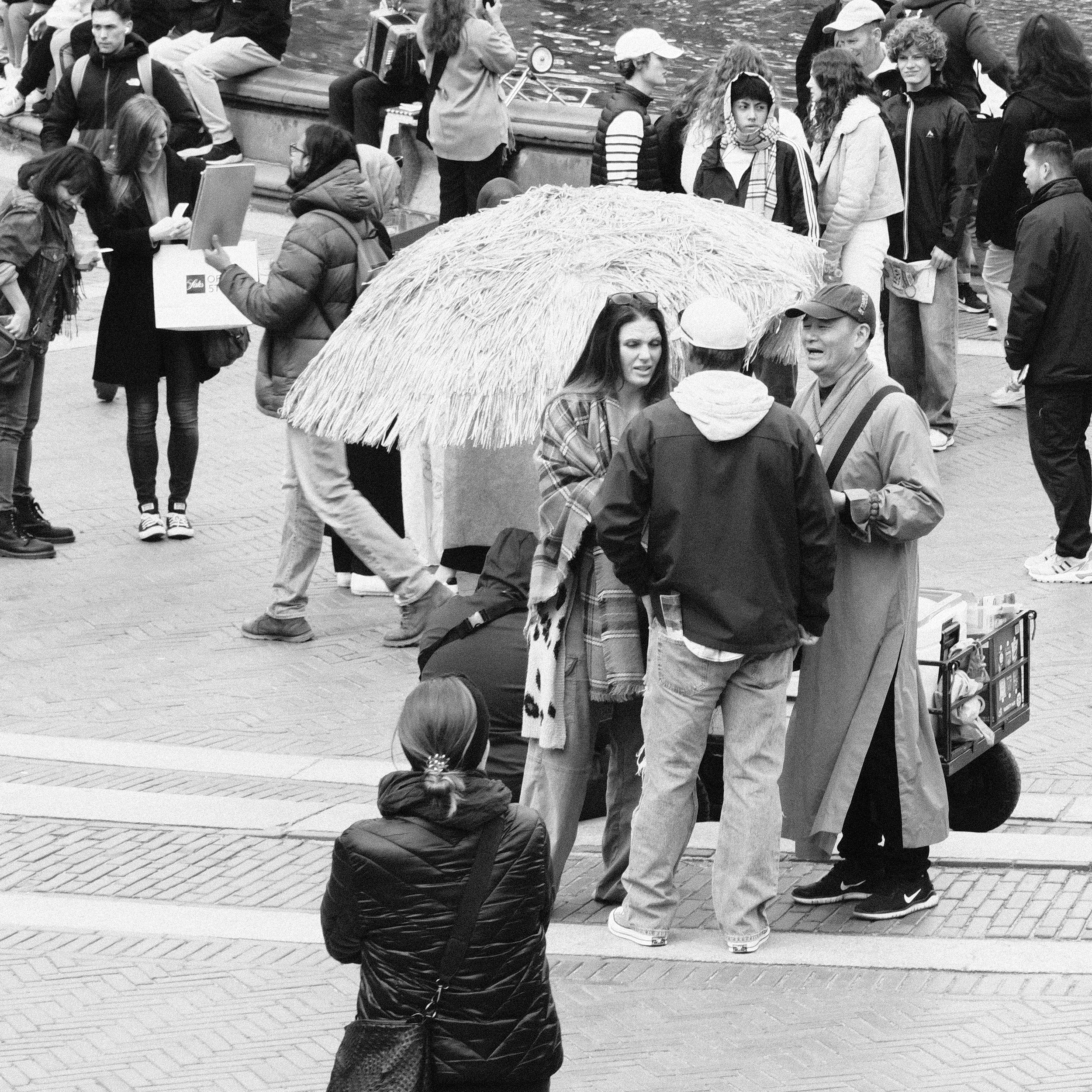 A group of people gathered on a city street, some sitting near a water fountain, others standing and talking, with a woman seated on the pavement in the foreground.