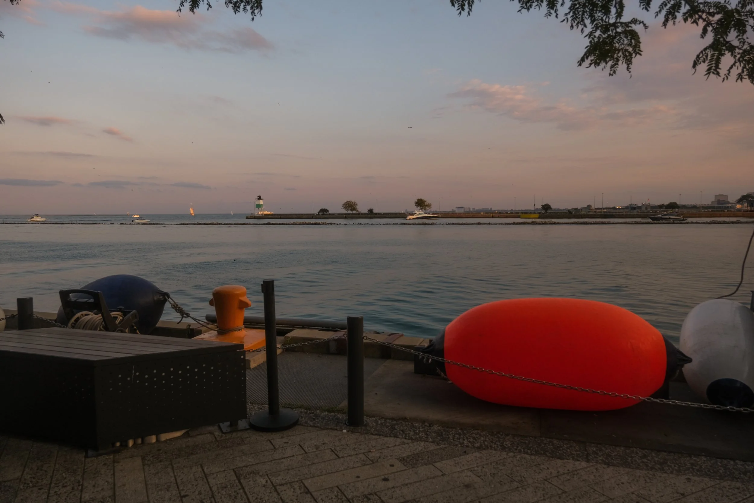 A waterfront scene during sunset with boats in the distance, a lighthouse on a breakwater, and large colorful buoys along the pier.