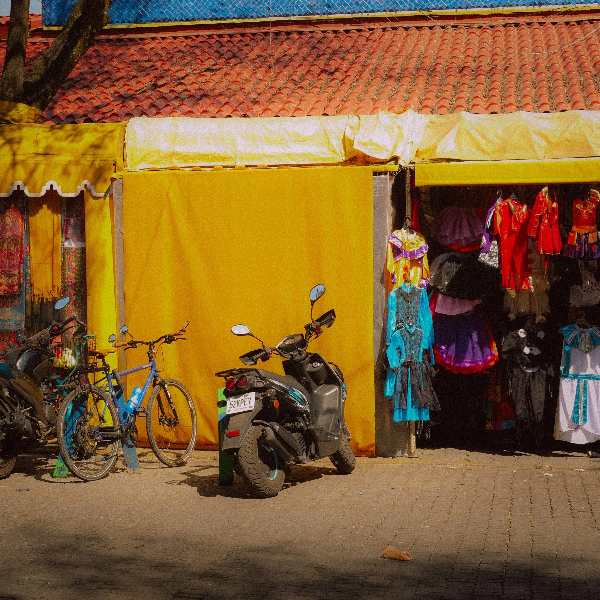A street scene with a yellow tented market stall selling colorful costumes and dresses. Two bicycles and a scooter are parked in front of the stall on a cobblestone pavement. The background features a building with a red tiled roof.