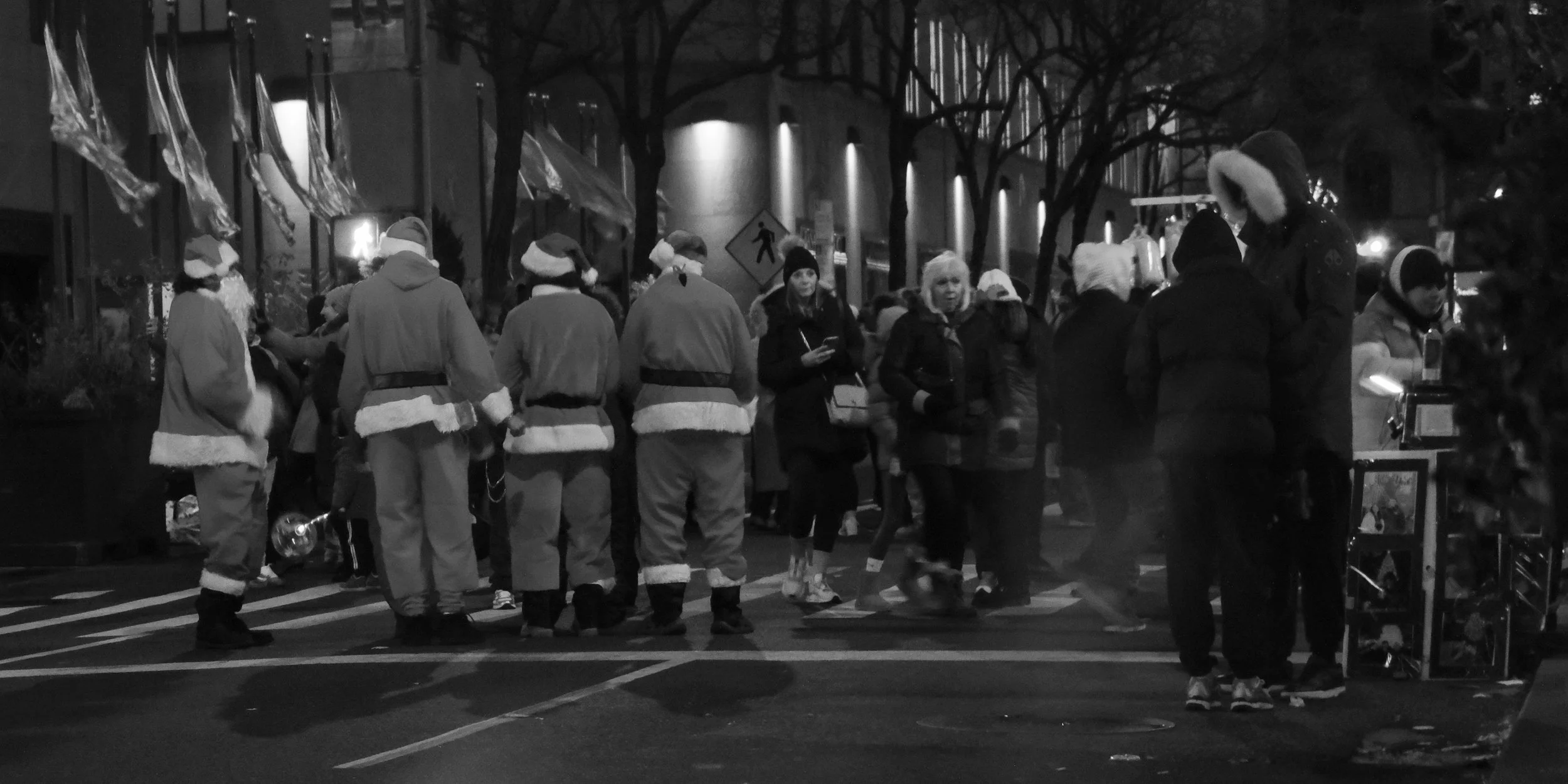People dressed in Santa costumes and winter clothes gather on a city street at night, some with Christmas hats, amid a festive, holiday-season atmosphere.
