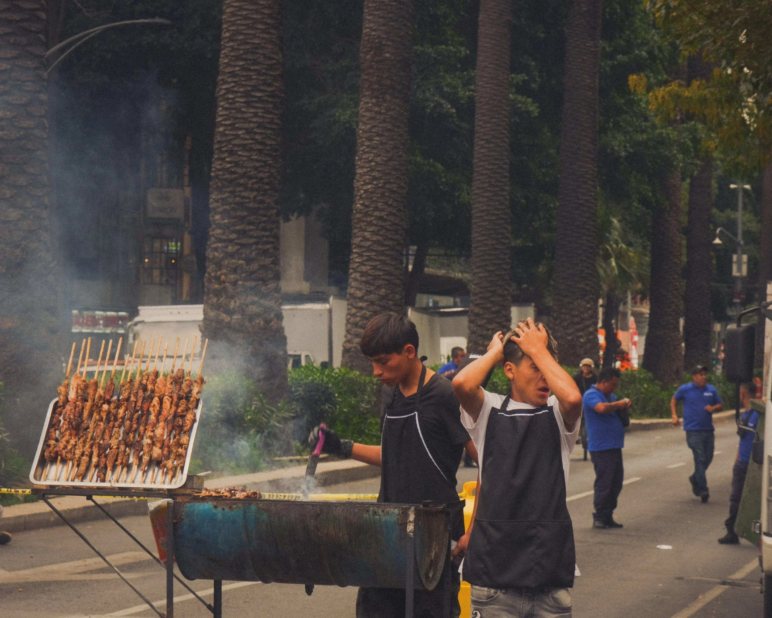 Two boys working at a street food stall with skewered meat, in an outdoor urban area with palm trees and several people in blue uniforms in the background.