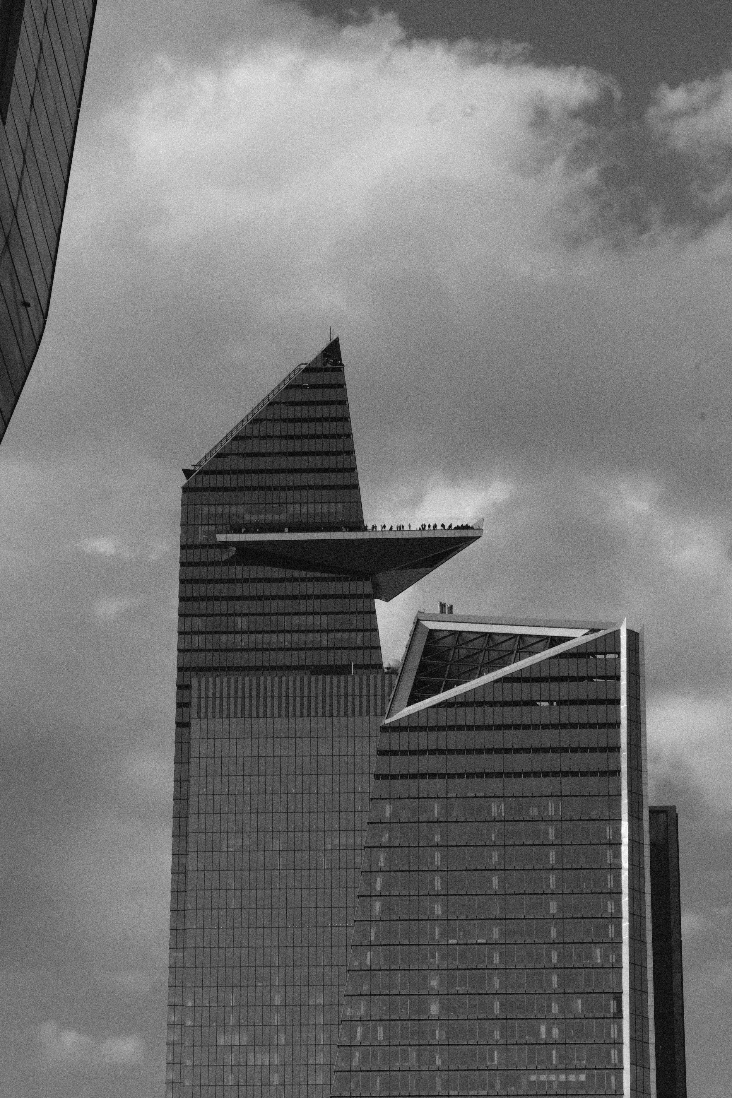 Black and white photo of a modern skyscraper with an overhanging section, with other tall buildings in the background and a cloudy sky.