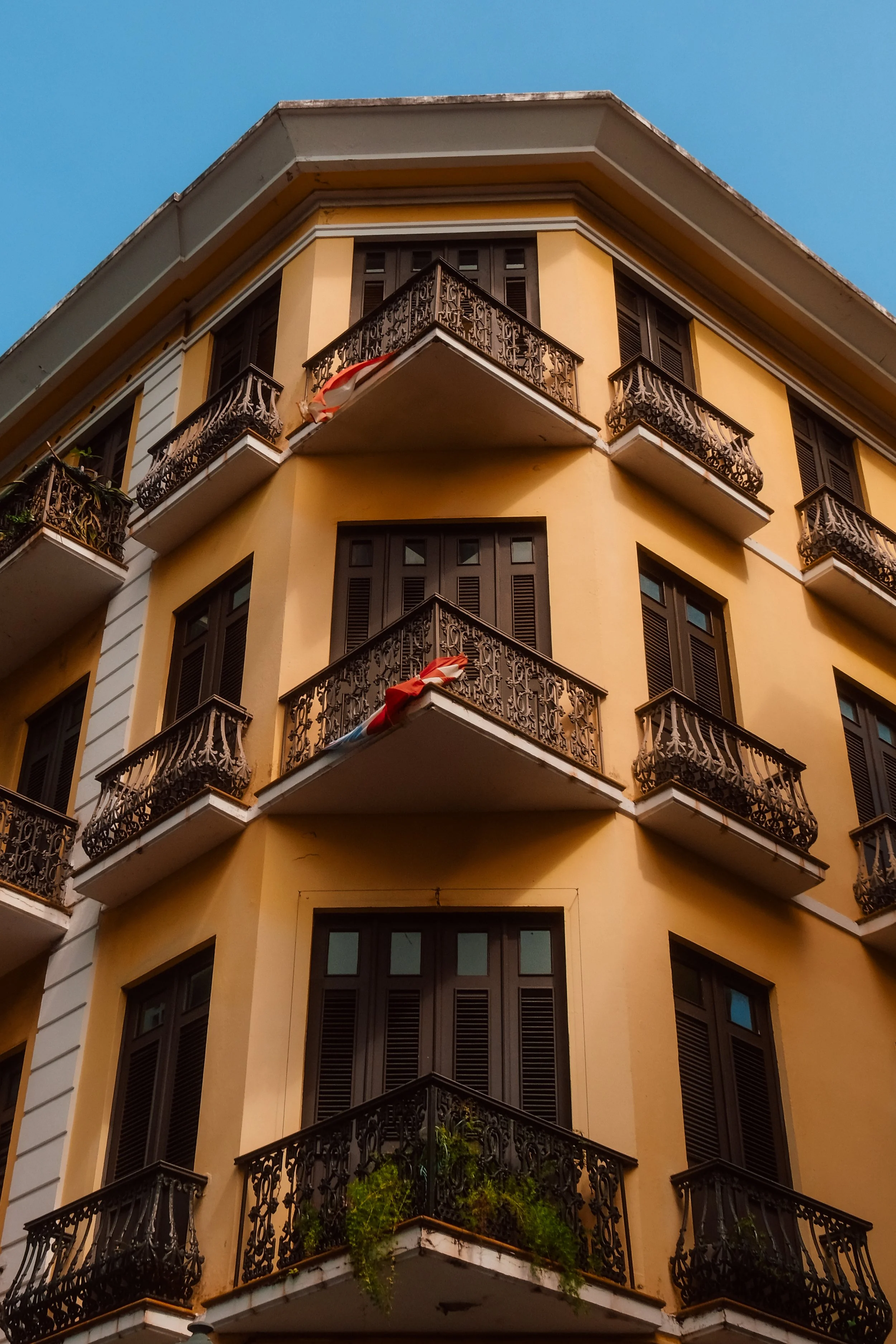 A yellow residential building with balconies having ornate black wrought iron railings, some with plants, under a clear blue sky.