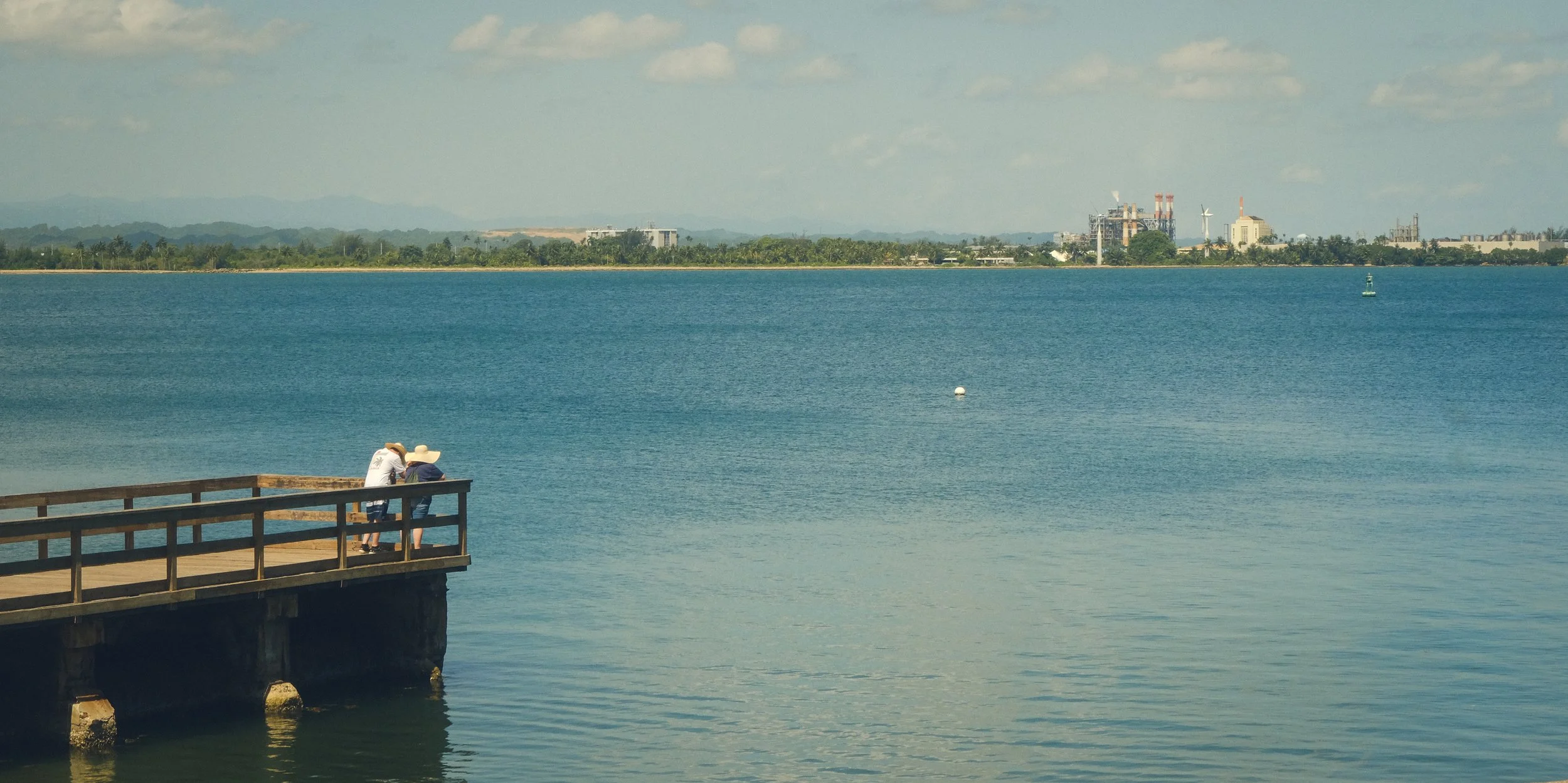 Two people stand on a wooden pier by the water, wearing large sun hats, with a large body of water and a distant industrial city skyline in the background.
