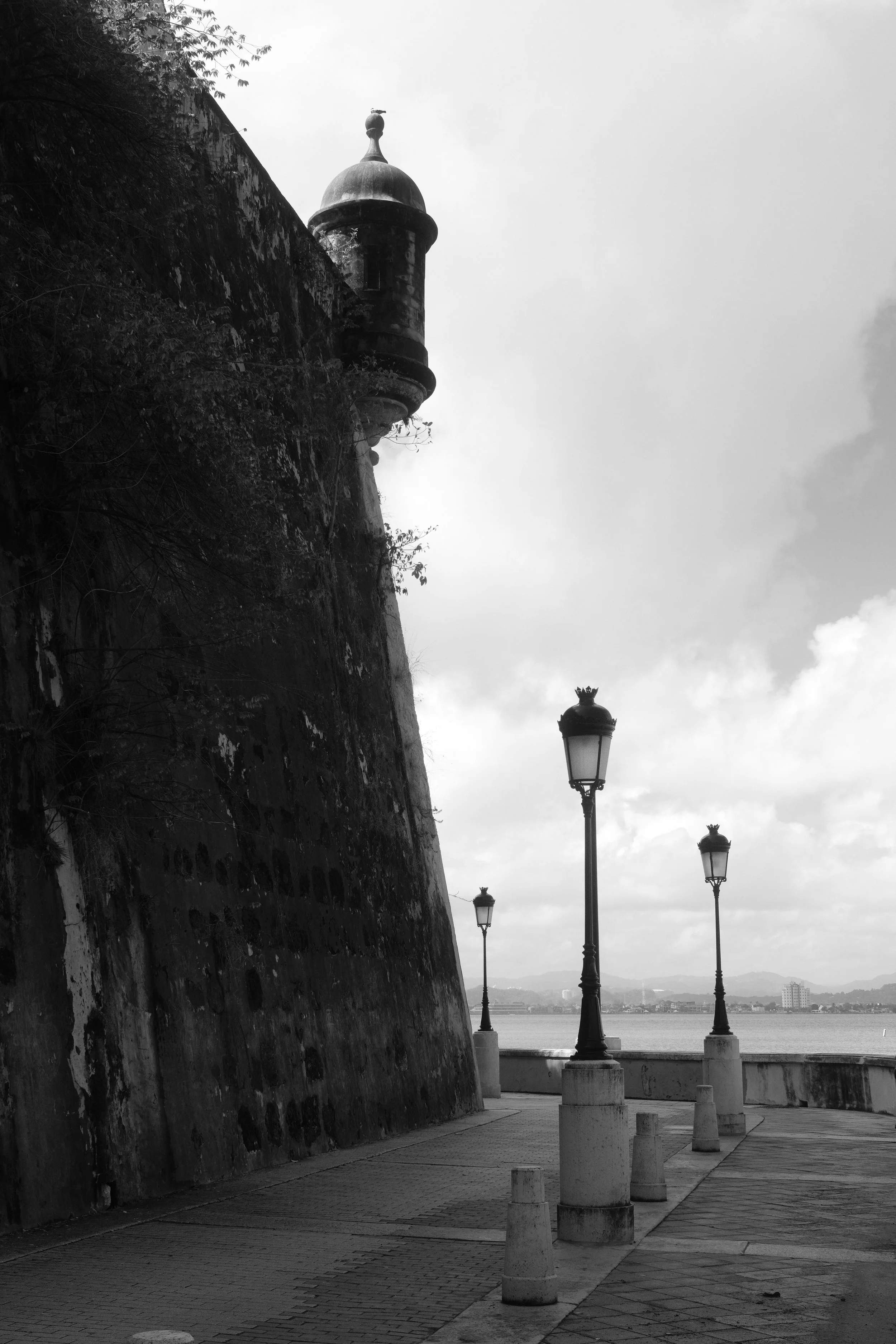 Black and white photo of a waterfront promenade with vintage-style street lamps, a large stone fortification wall, and a distant city skyline with hills in the background.