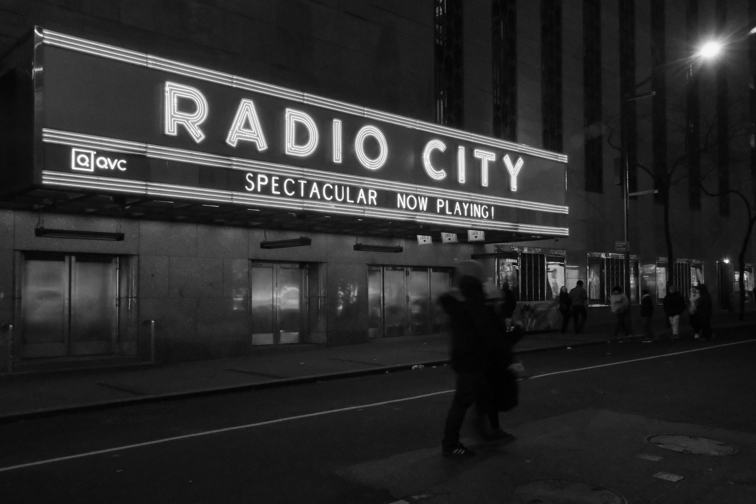Nighttime scene of Radio City music hall with a lit marquee displaying the words 'RADIO CITY' and 'SPECTACULAR NOW PLAYING!', with pedestrians walking on the sidewalk.