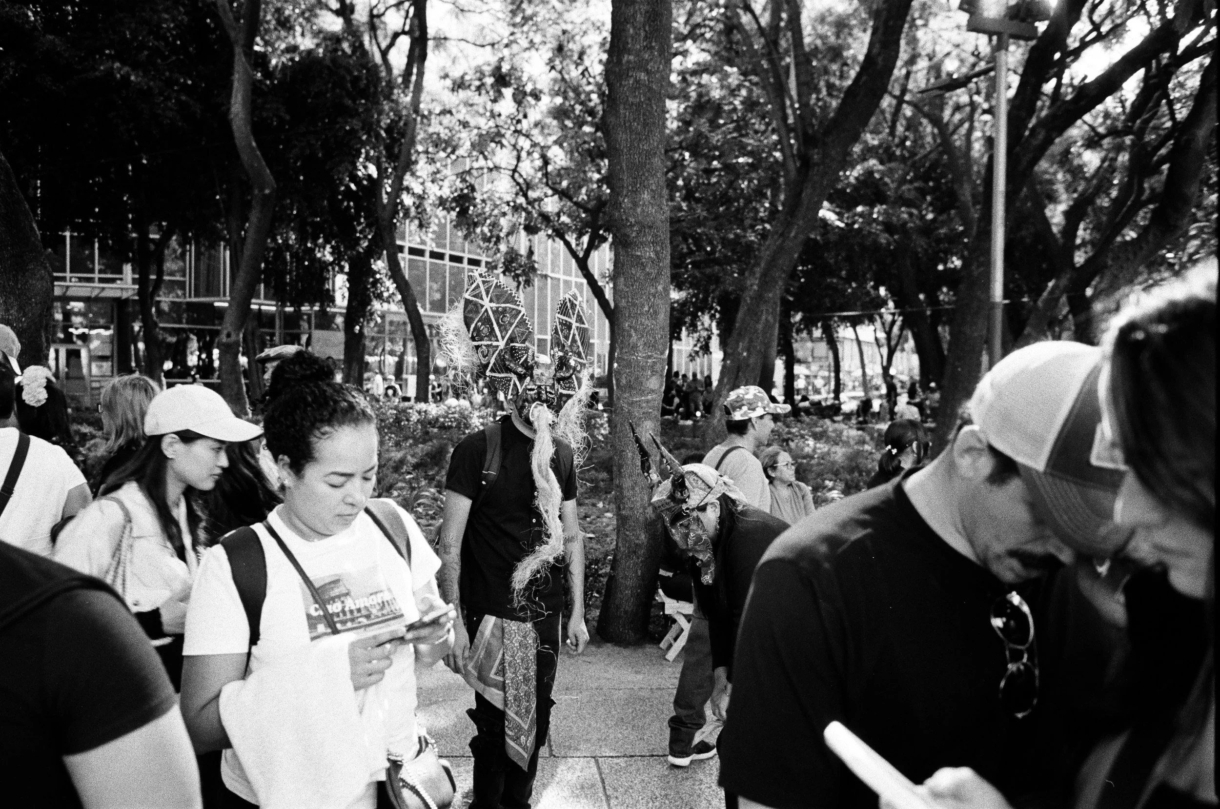 People walking outdoors, some wearing colorful masks and costumes, in a park with trees and a building in the background.