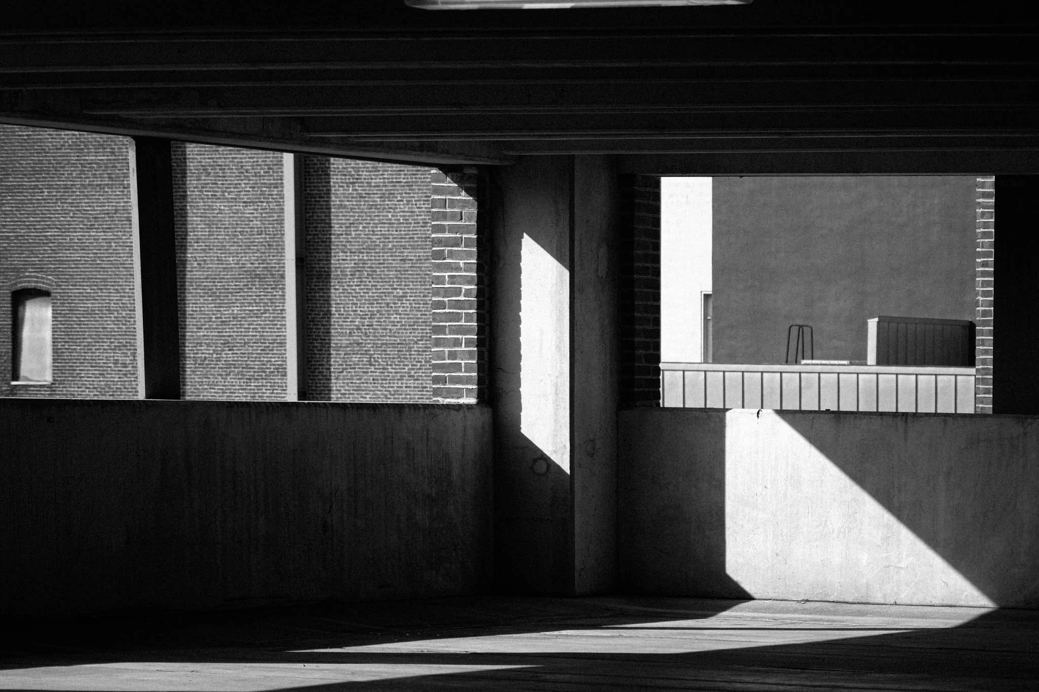Black and white photo of a building's exterior showing walls made of brick and concrete, with strong shadows cast by sunlight on the surfaces, and a distant rooftop structure visible.