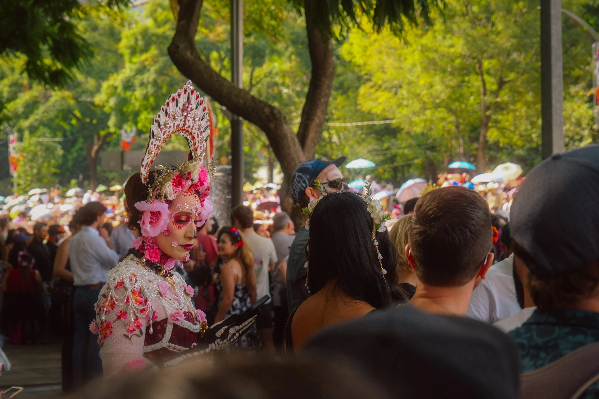 Person dressed in an elaborate costume with makeup and floral headpiece, participating in a cultural event or parade among a crowd outdoors in a park with trees.