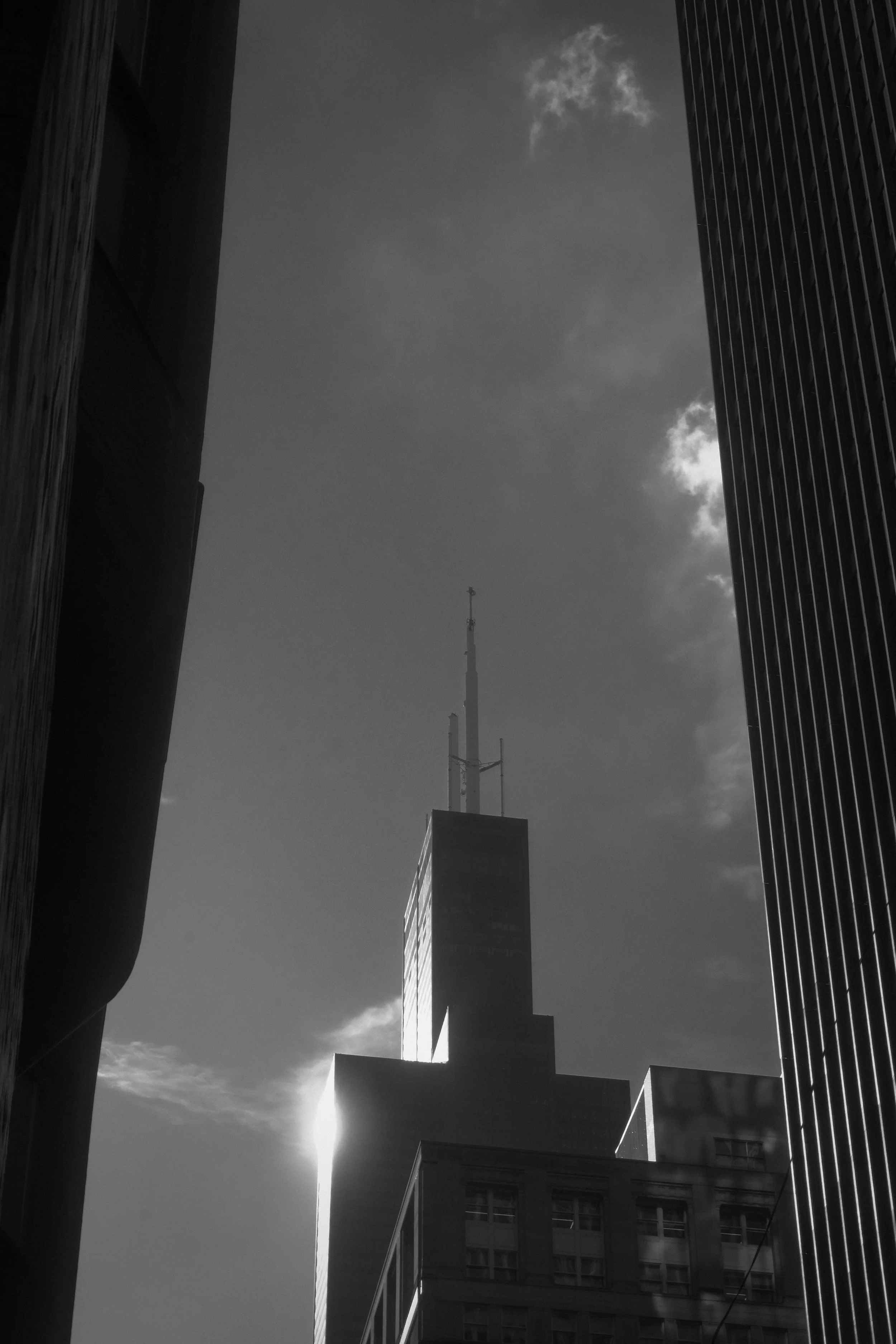 Black and white photo of a skyscraper with antennas on top, surrounded by other tall buildings, with the sun setting behind it.