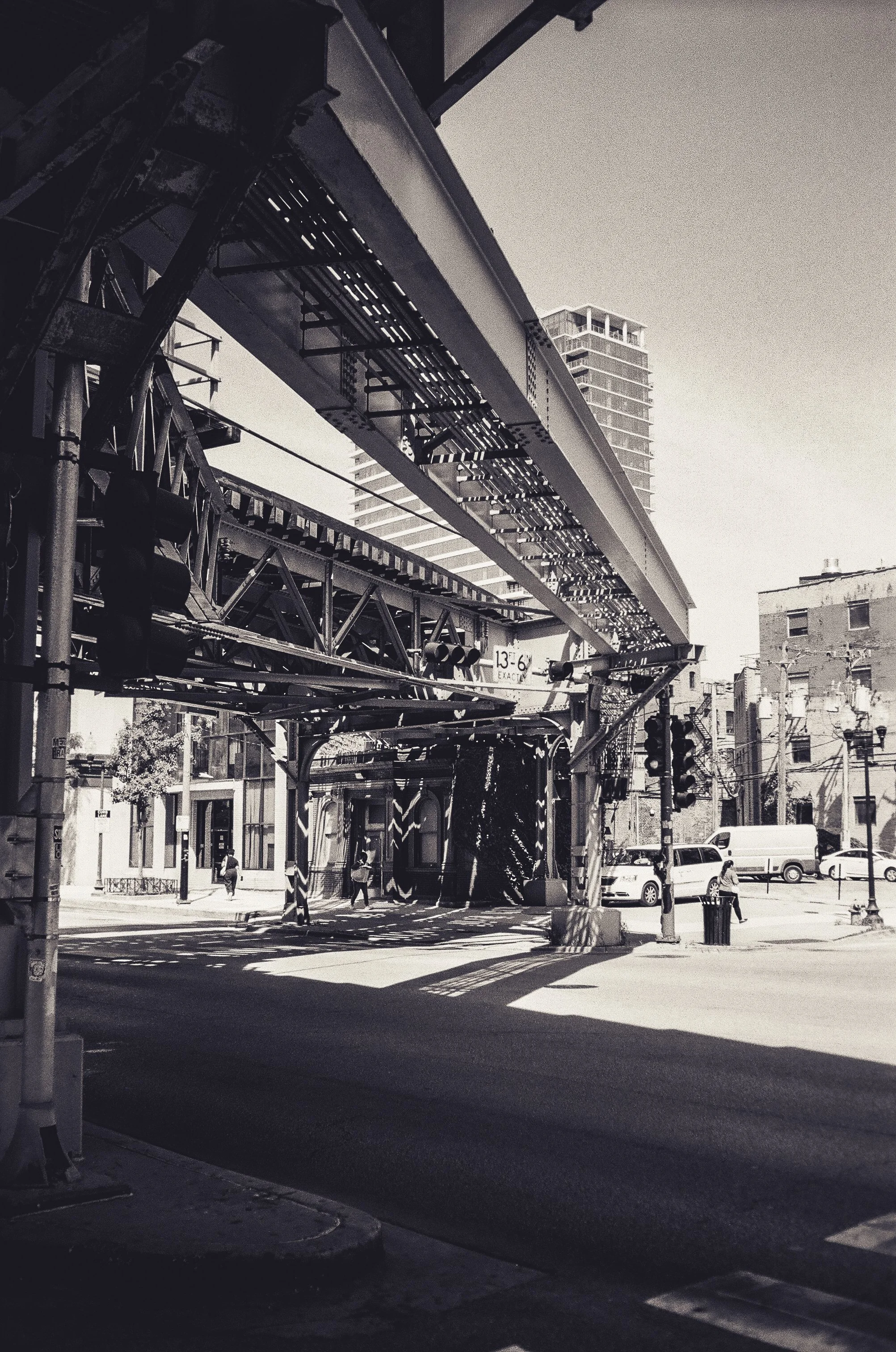 Black and white photo of an urban street scene with elevated train tracks, traffic lights, and pedestrians walking on the sidewalk. Tall buildings are visible in the background.