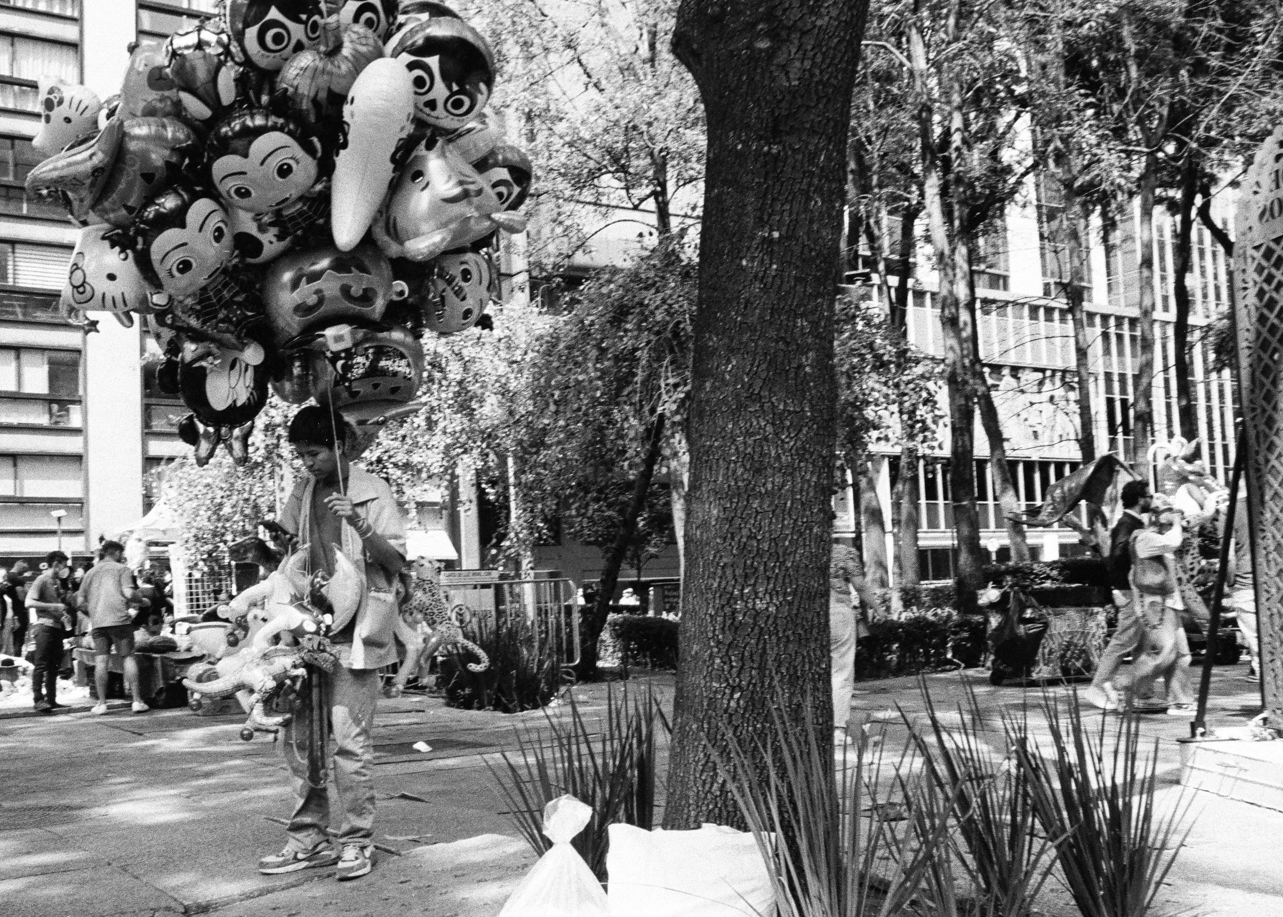 A person holding a large bunch of cartoon character balloons, standing on a city sidewalk near a tree, with others walking and vendor stalls in the background.