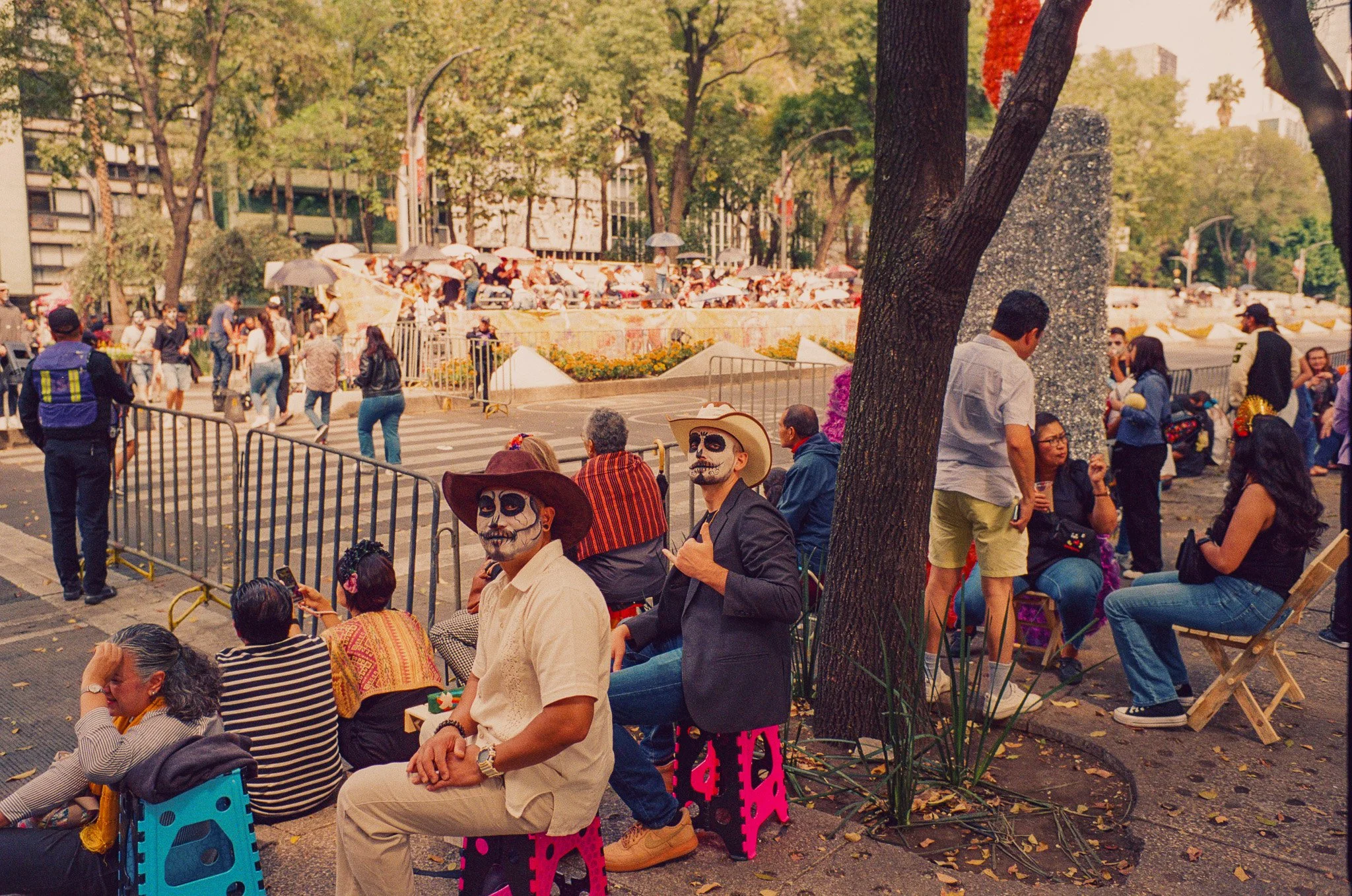 Crowd gathering with some people wearing Día de los Muertos themed face paint and costumes in a park with trees and city buildings in the background.