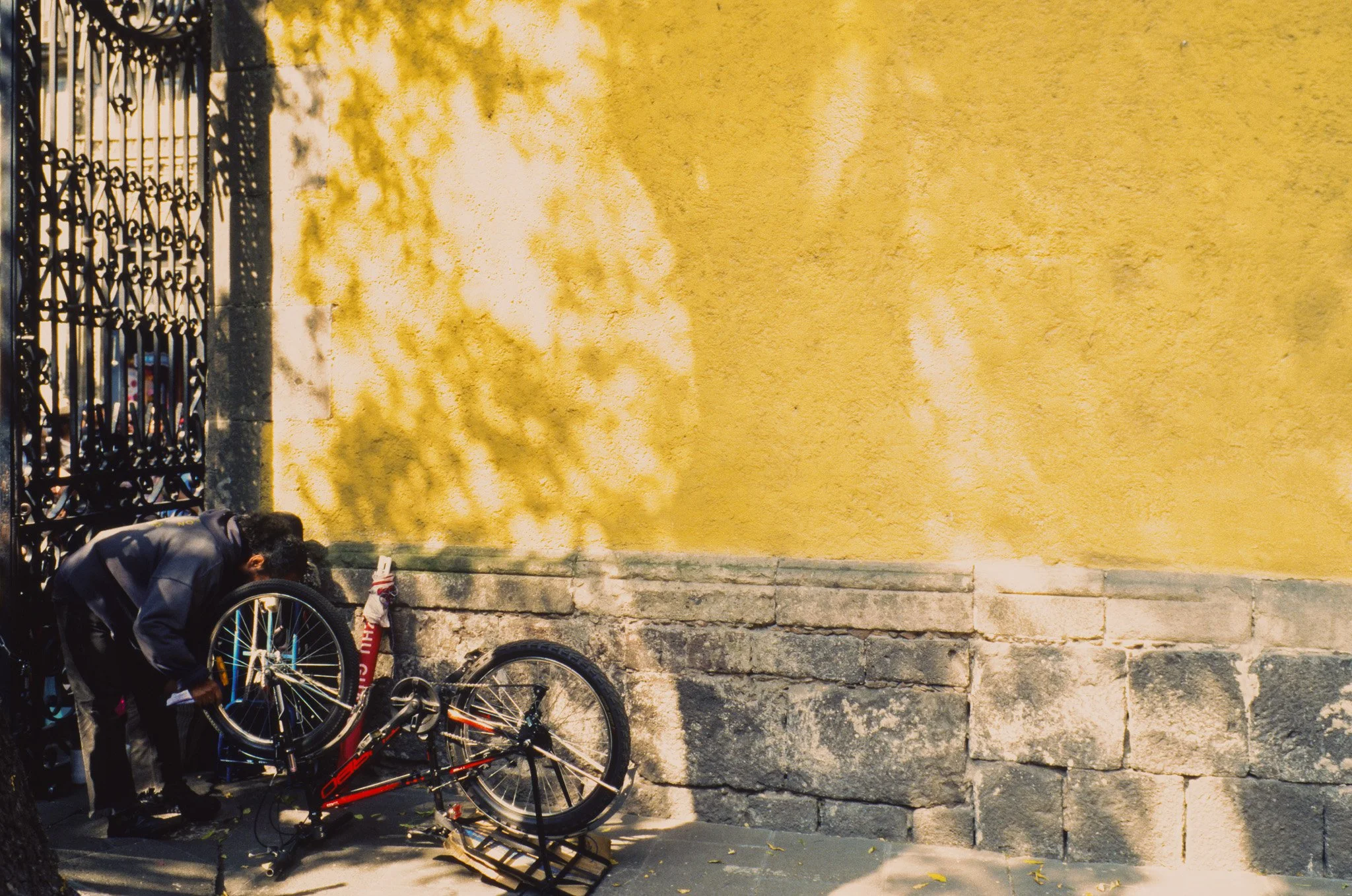 A person is repairing a red bicycle against a yellow wall with shadows of leaves. The person is bending over, working on the bicycle on the sidewalk.