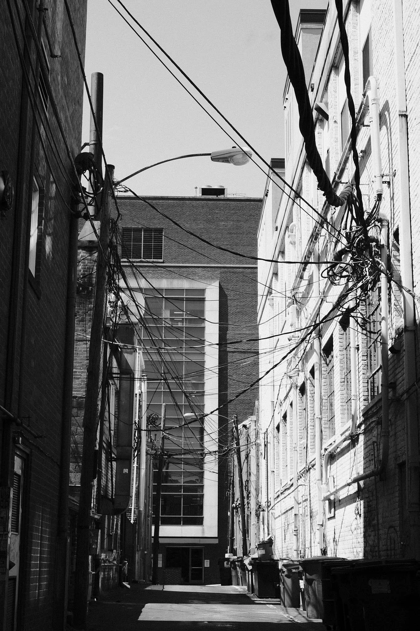 An alleyway with tangled overhead power lines, brick buildings on either side, and a modern building with large windows at the end.