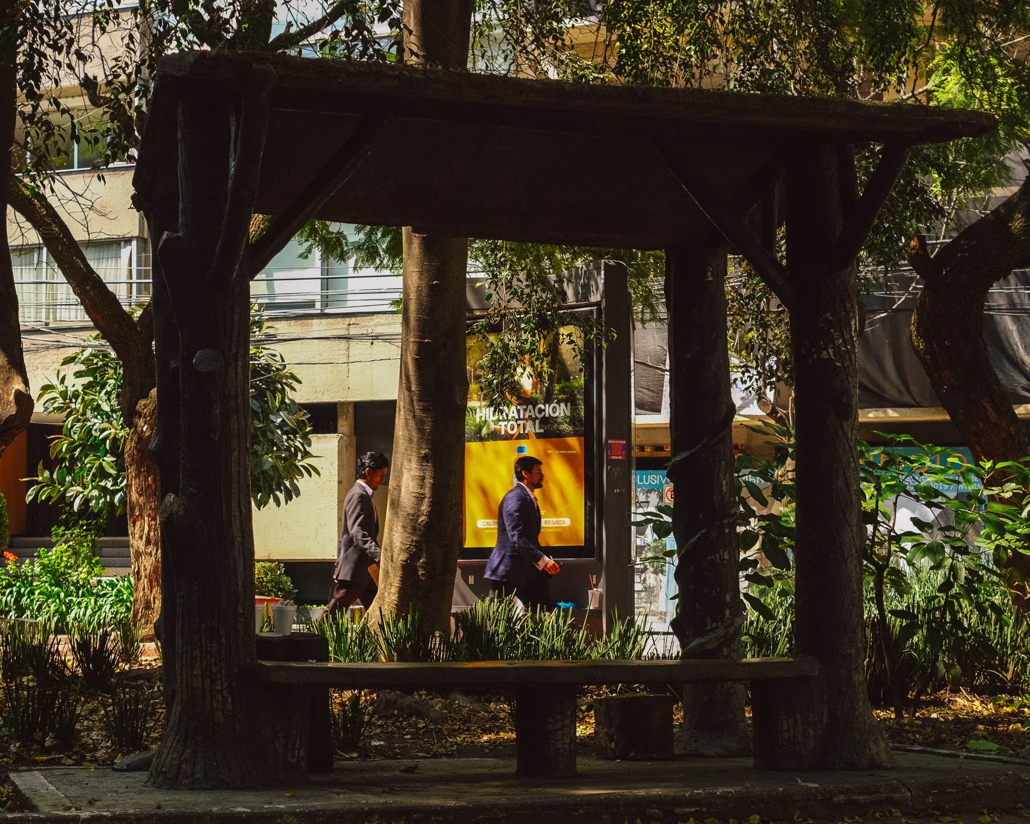 A park bench surrounded by trees with two men walking by in business attire, a glowing advertisement billboard in the background, and urban buildings behind.