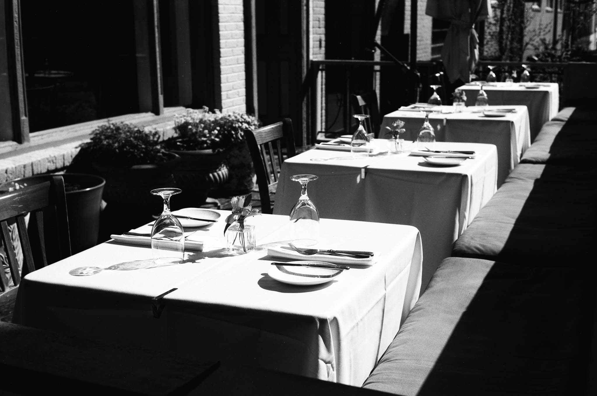 Empty outdoor restaurant tables with white tablecloths, upside-down glasses, plates, and silverware, shaded by sunlight on a patio with brick wall and windows.