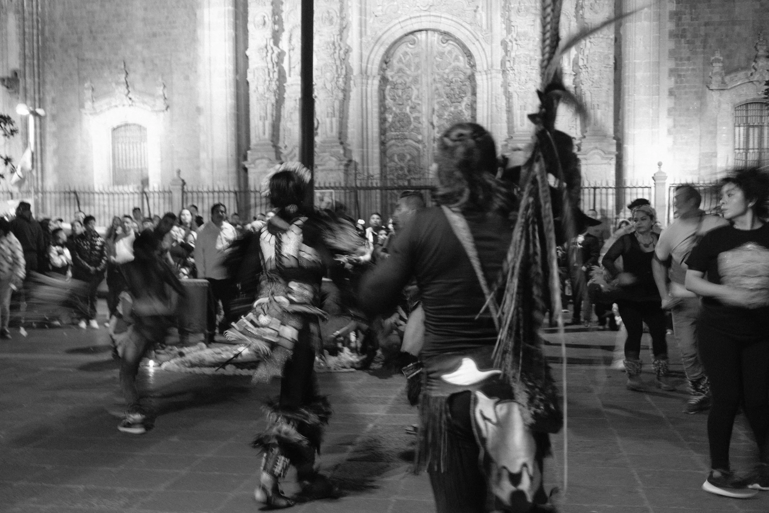People gathered in a plaza at night, with some dancing and others watching, in front of a historic building with ornate architecture.