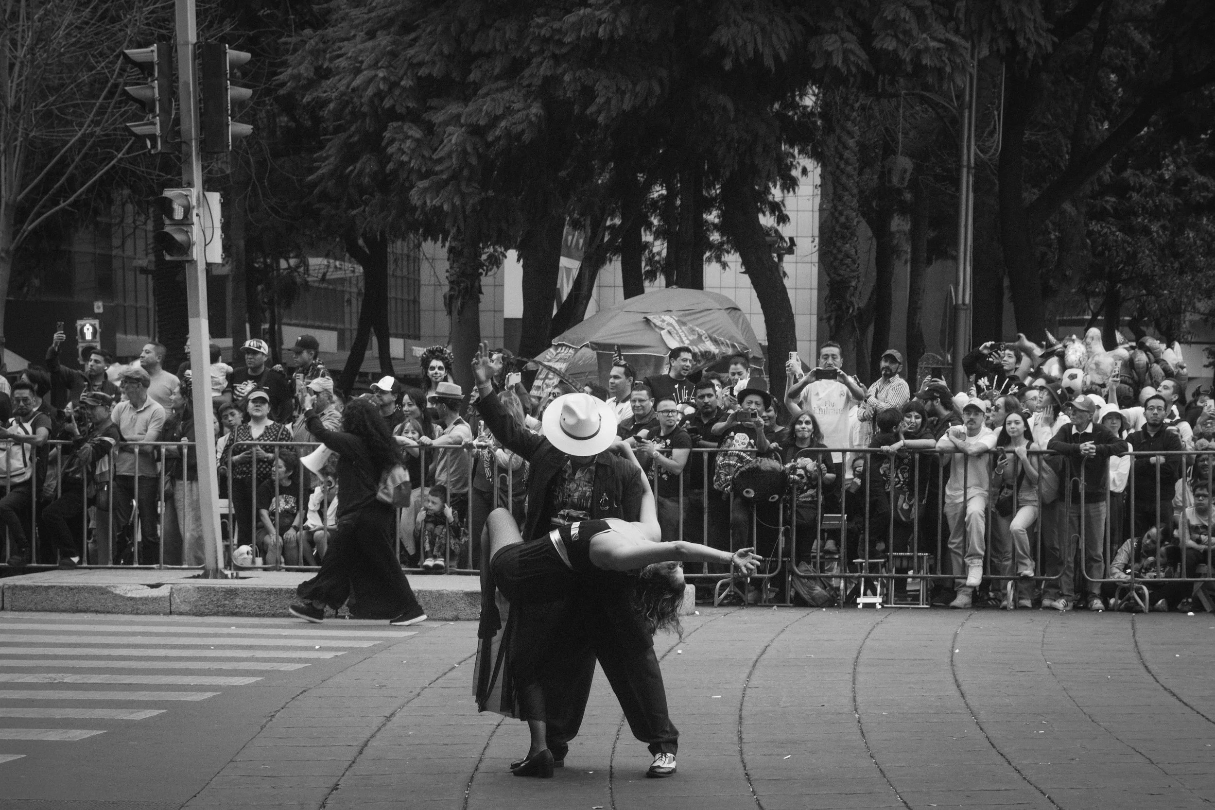 Street performance with a solo dancer executing a lift, crowd behind barriers watching, some taking photos, daytime outdoor. Black and white.