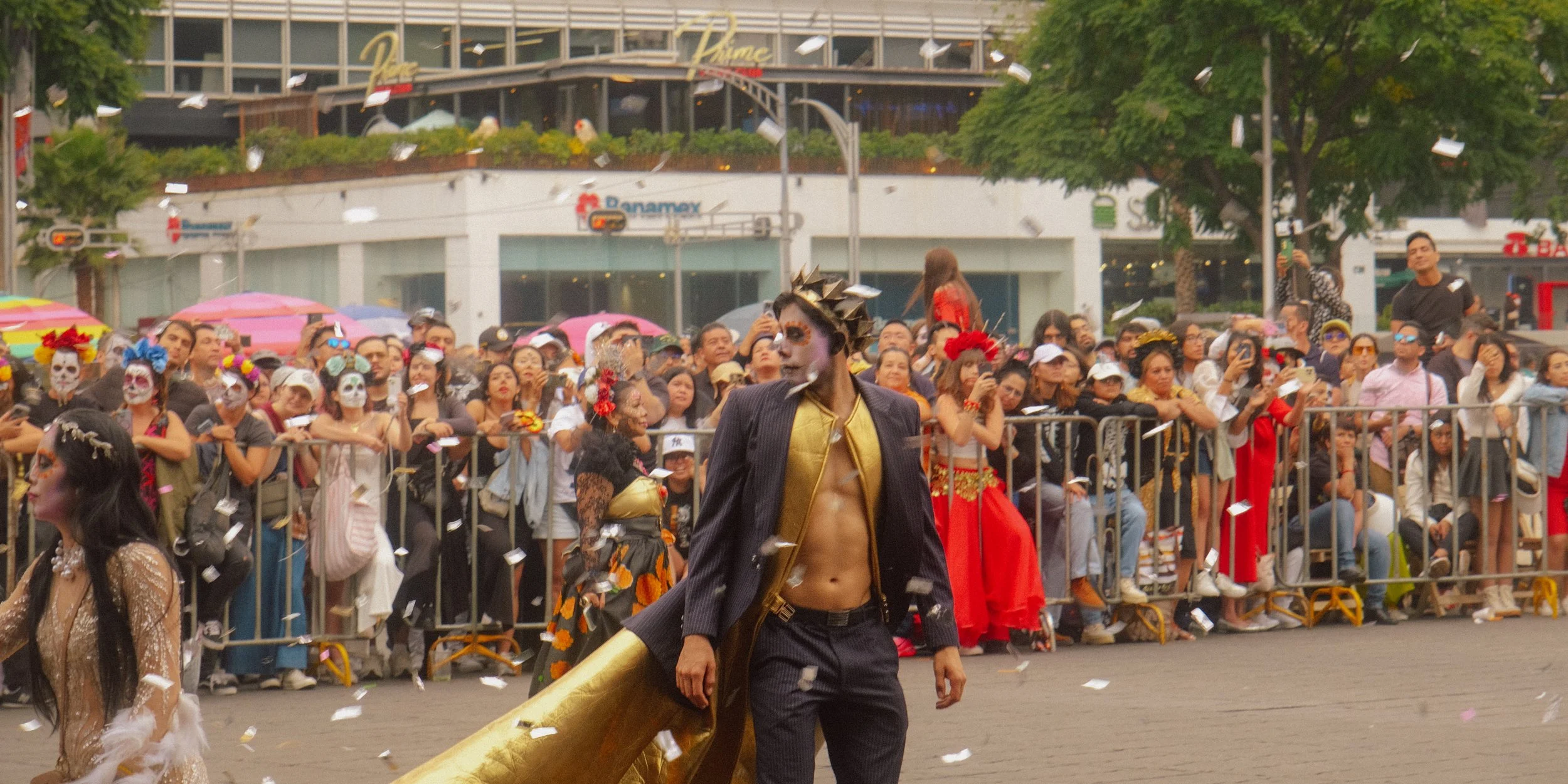 A person dressed as a skeleton with face paint, wearing a pinstripe suit and golden cape, standing in front of a crowd of spectators at a parade, some in costumes, some taking photos, with confetti falling.