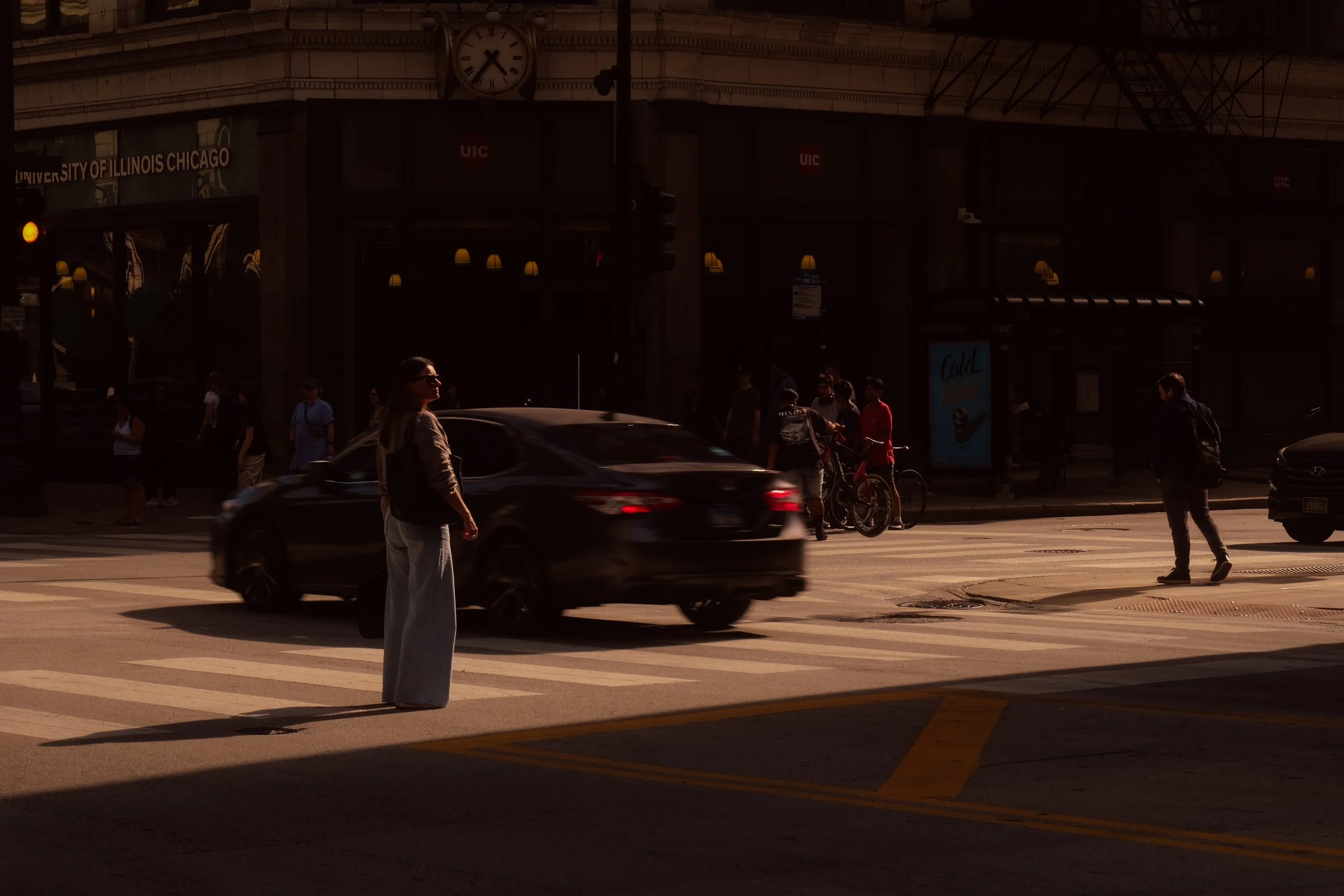 City street scene with pedestrians crossing the crosswalk, a woman standing while waiting to cross, a black car driving through, and people in the background near a bus stop and building labeled 'University of Illinois Chicago'.