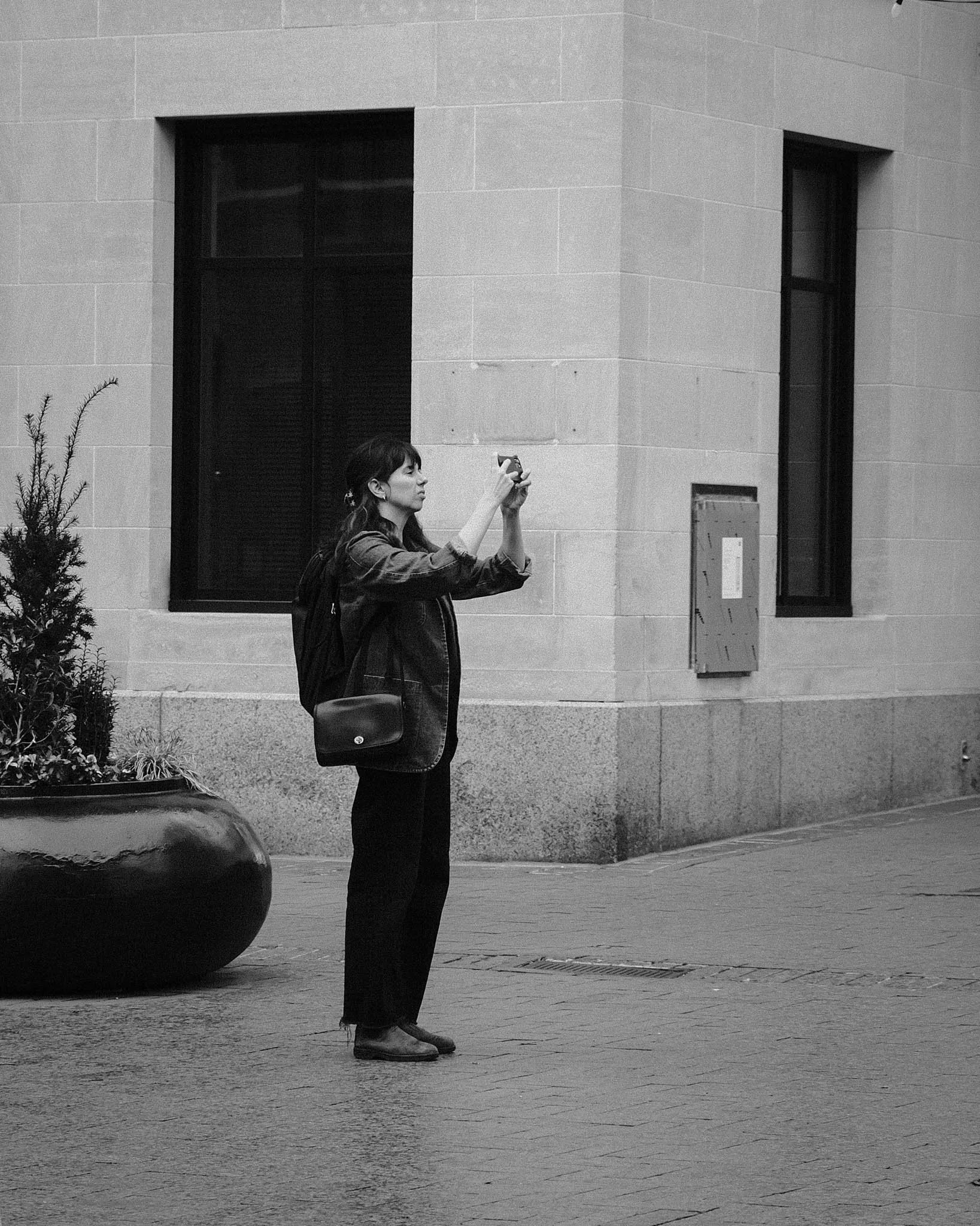 A woman taking a photo with her phone on a city sidewalk in black and white.
