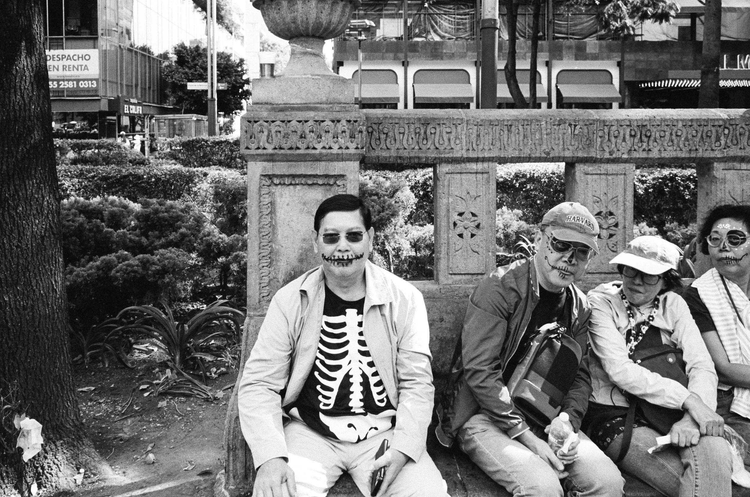 Four people sitting on a park bench, wearing skeleton face paint and sunglasses, with trees and a decorative stone structure behind them.