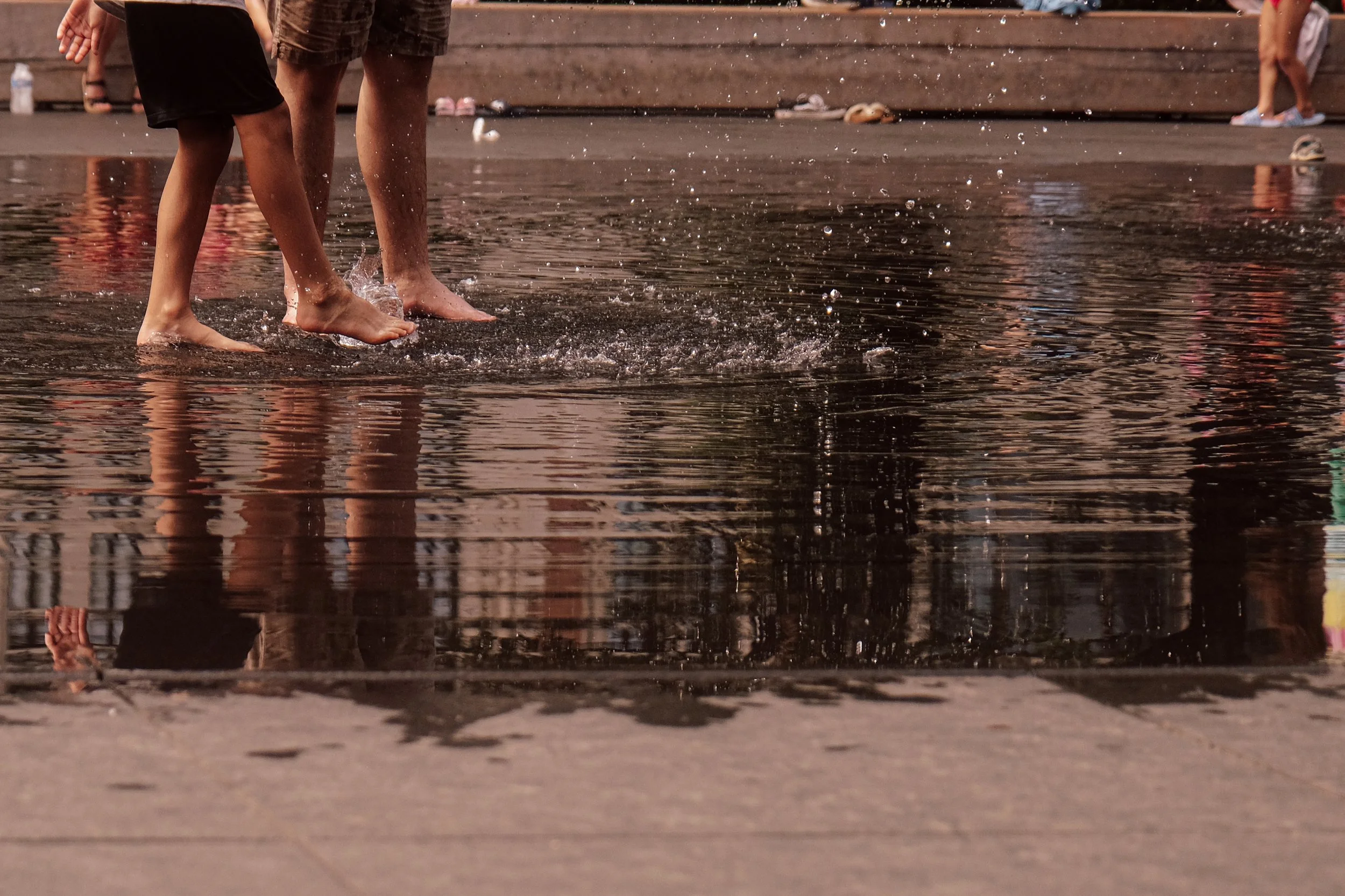 Close-up of two children walking barefoot through a shallow puddle, splashing water, with people and shoes visible in the background.