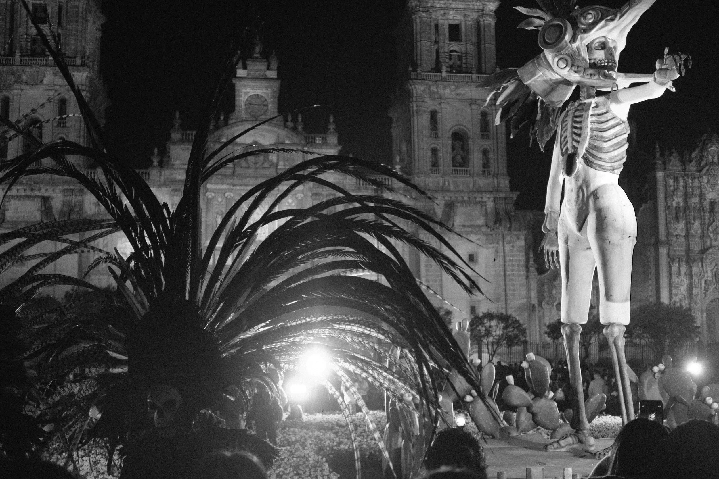 A black and white photo of a large skeletal figure with a mask on its face, standing in front of a historical building at night. The figure has an elaborate headpiece, and there are people gathered nearby.