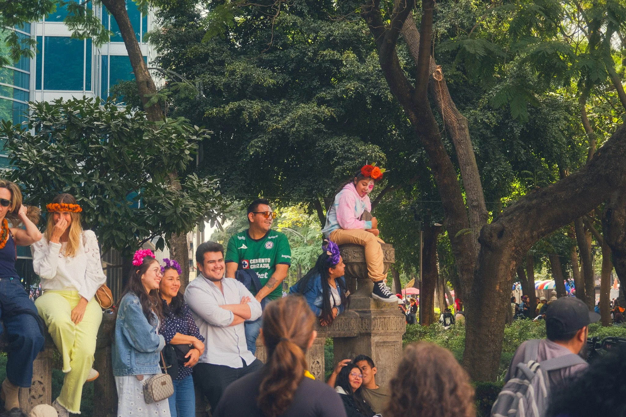 Group of people outdoors with some wearing flower crowns, smiling and looking towards a woman dressed as a clown sitting on a stone ledge, in a park with trees and a city building in the background.