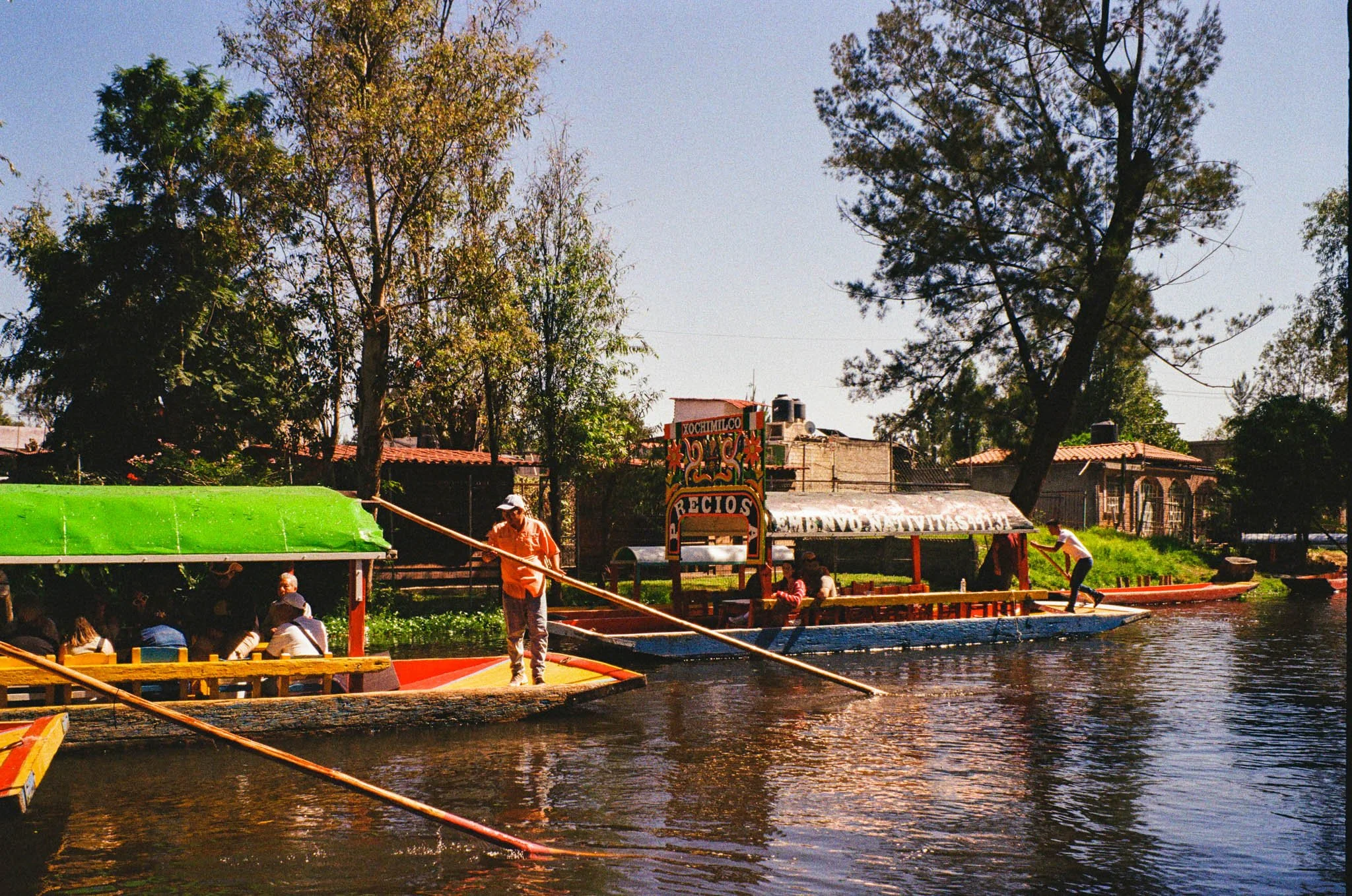 People on traditional Mexican floating platforms on a river, surrounded by trees and houses, with a clear blue sky.
