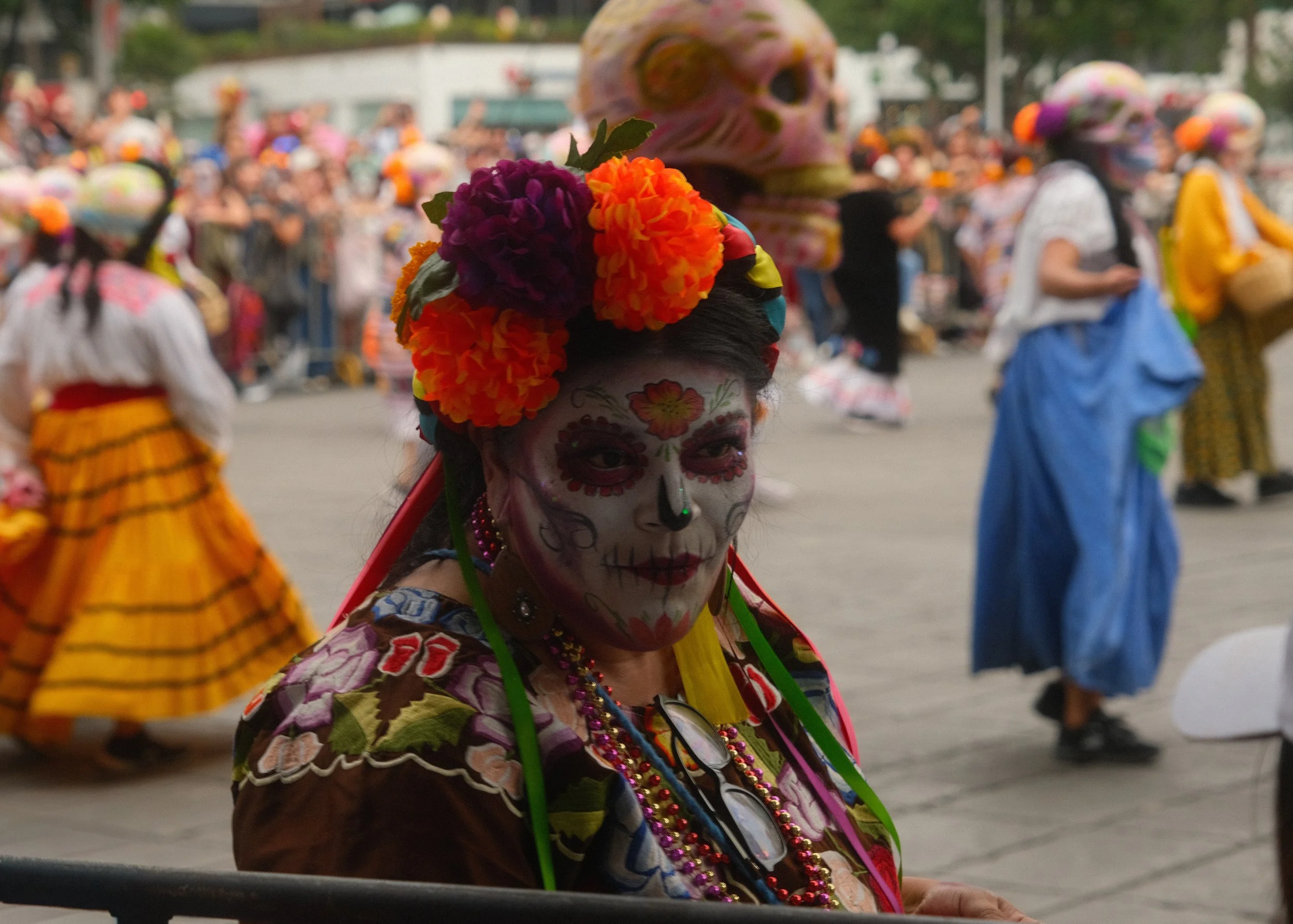 Woman with traditional Day of the Dead face paint, wearing a flower crown of orange, purple, and pink flowers, colorful clothing with floral embroidery, and multiple necklaces, standing among dancers dressed in colorful costumes at a cultural celebra