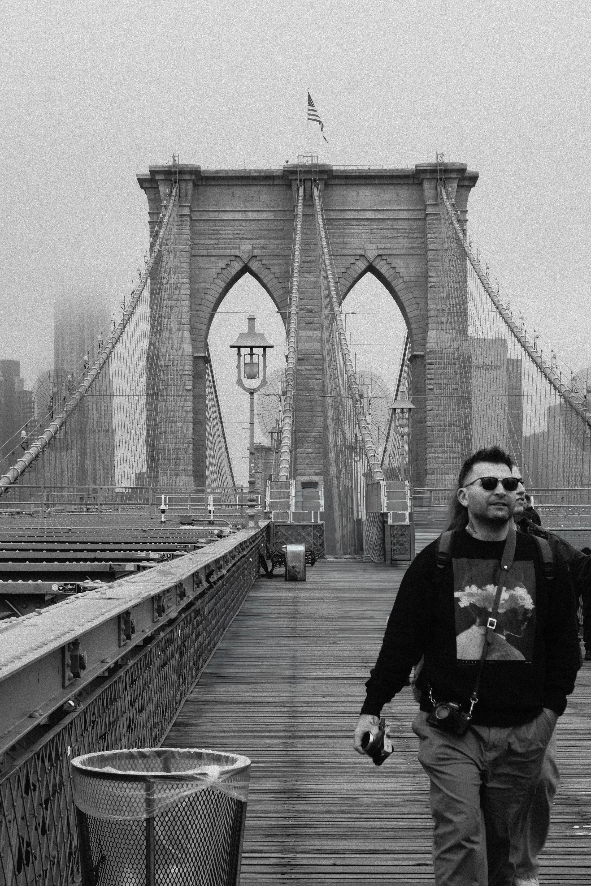 Black and white photo of Brooklyn Bridge in New York City with a man wearing sunglasses, a backpack, and a camera walking on the bridge.