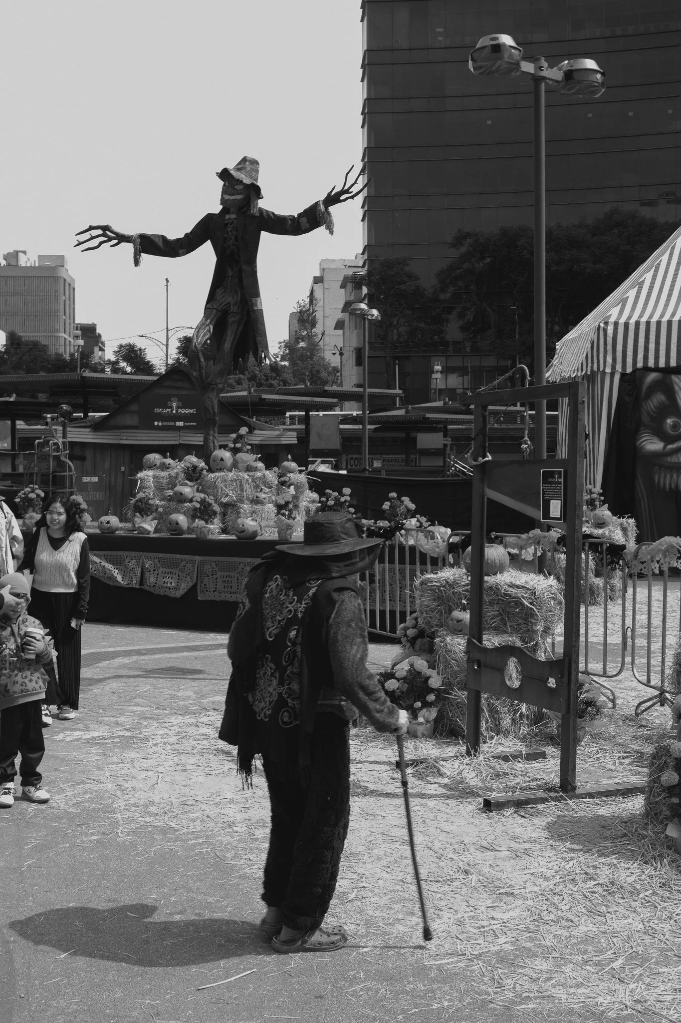 People walking in an outdoor festival or market with a large scarecrow or witch figure in the background, surrounded by pumpkins and hay bales.