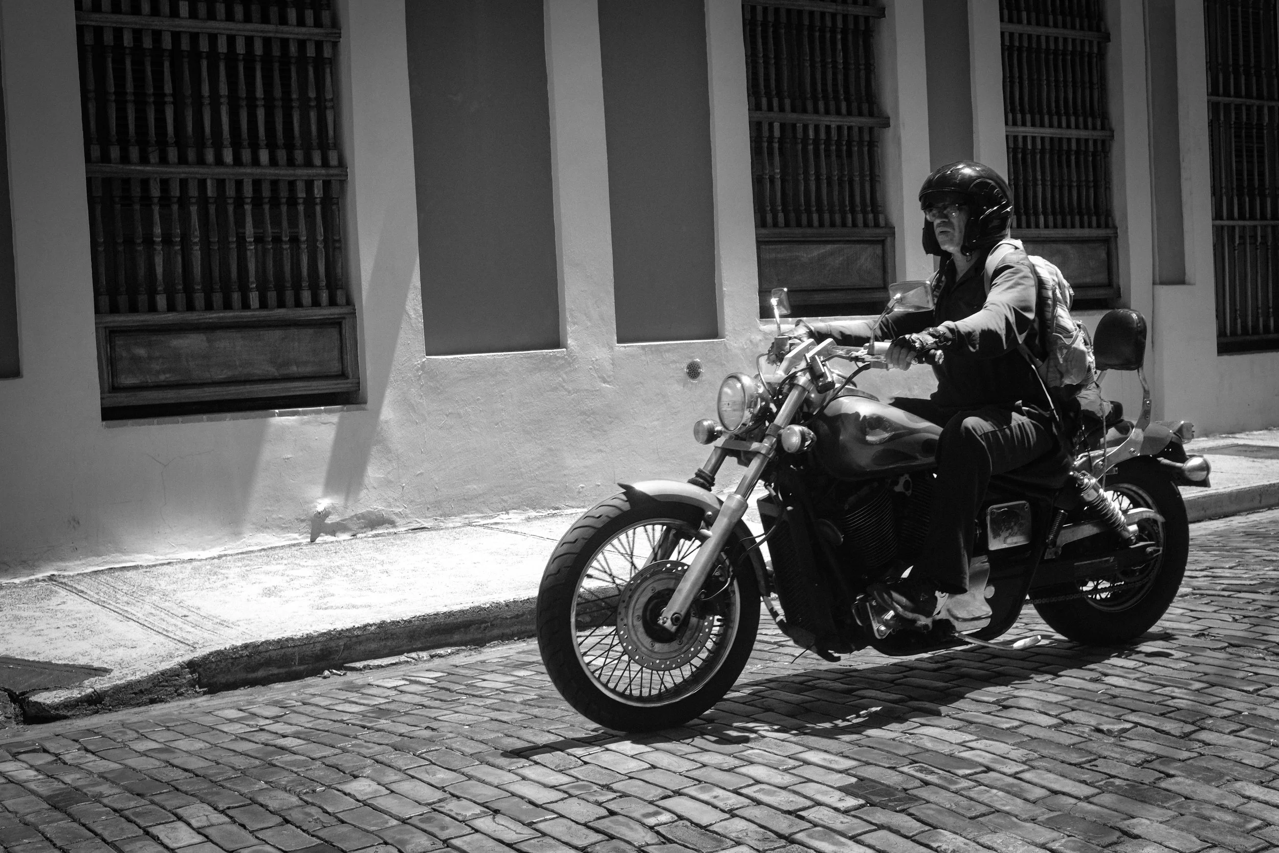 A person riding a motorcycle on a cobblestone street, wearing a helmet and backpack, with a building wall in the background.