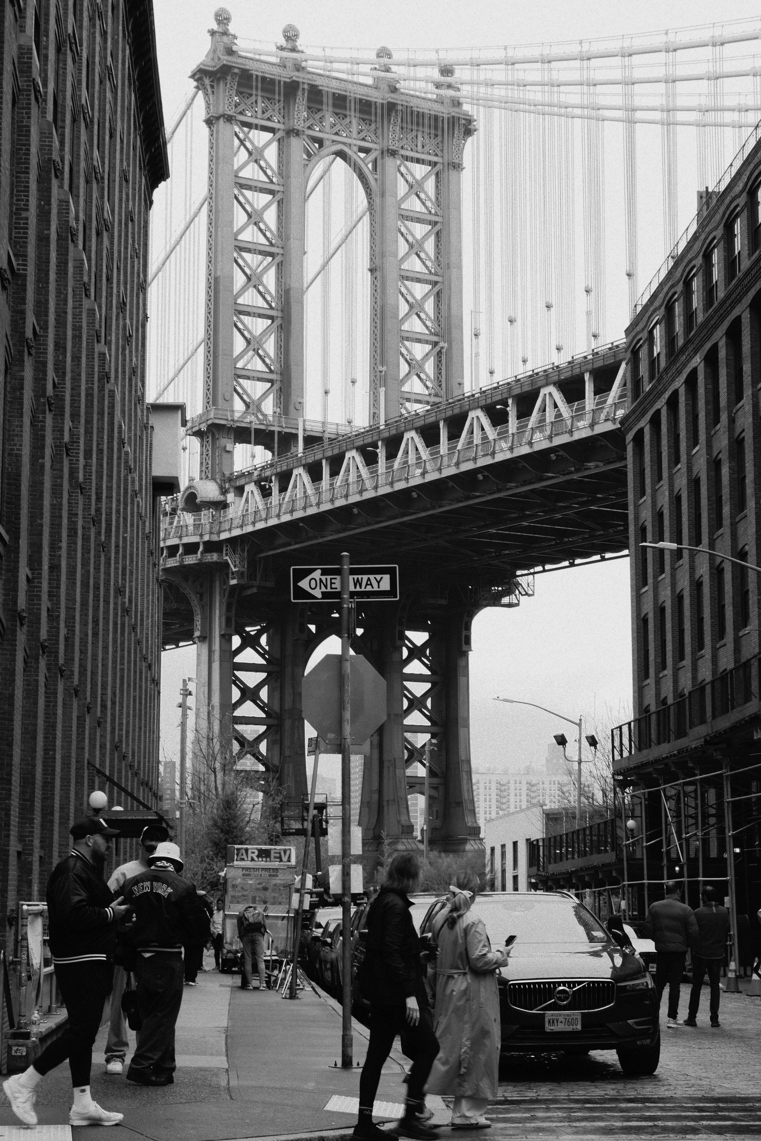Black and white photo of the Brooklyn Bridge in New York City viewed from a street with pedestrians and cars.