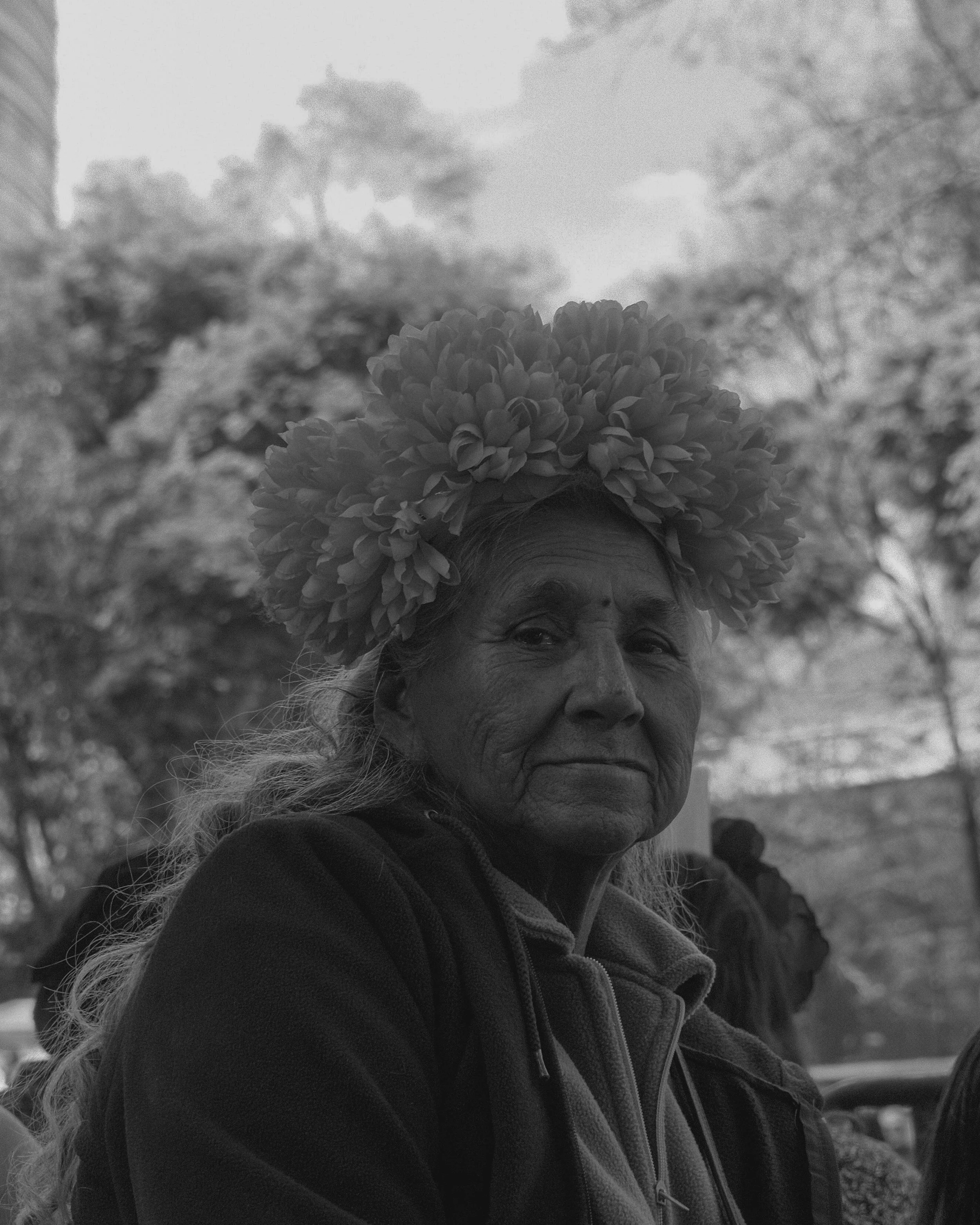 An elderly woman with a large flower crown on her head, outdoors with trees in the background.