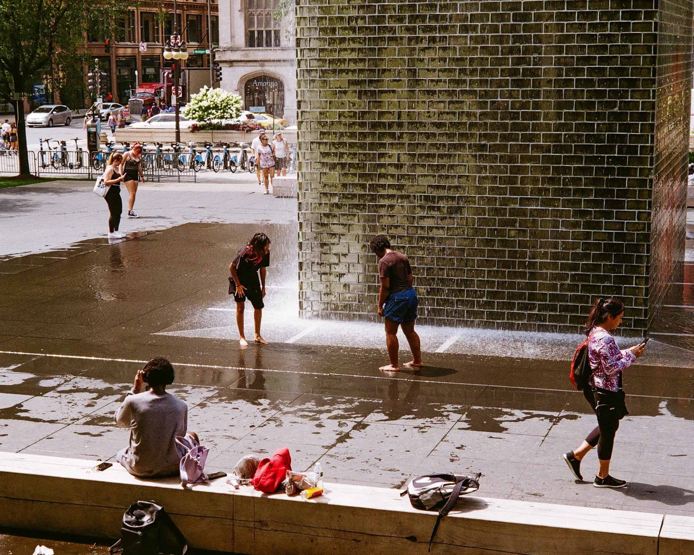 People enjoying a water fountain on a city sidewalk during the daytime, with some sitting and others walking or playing near the fountain.