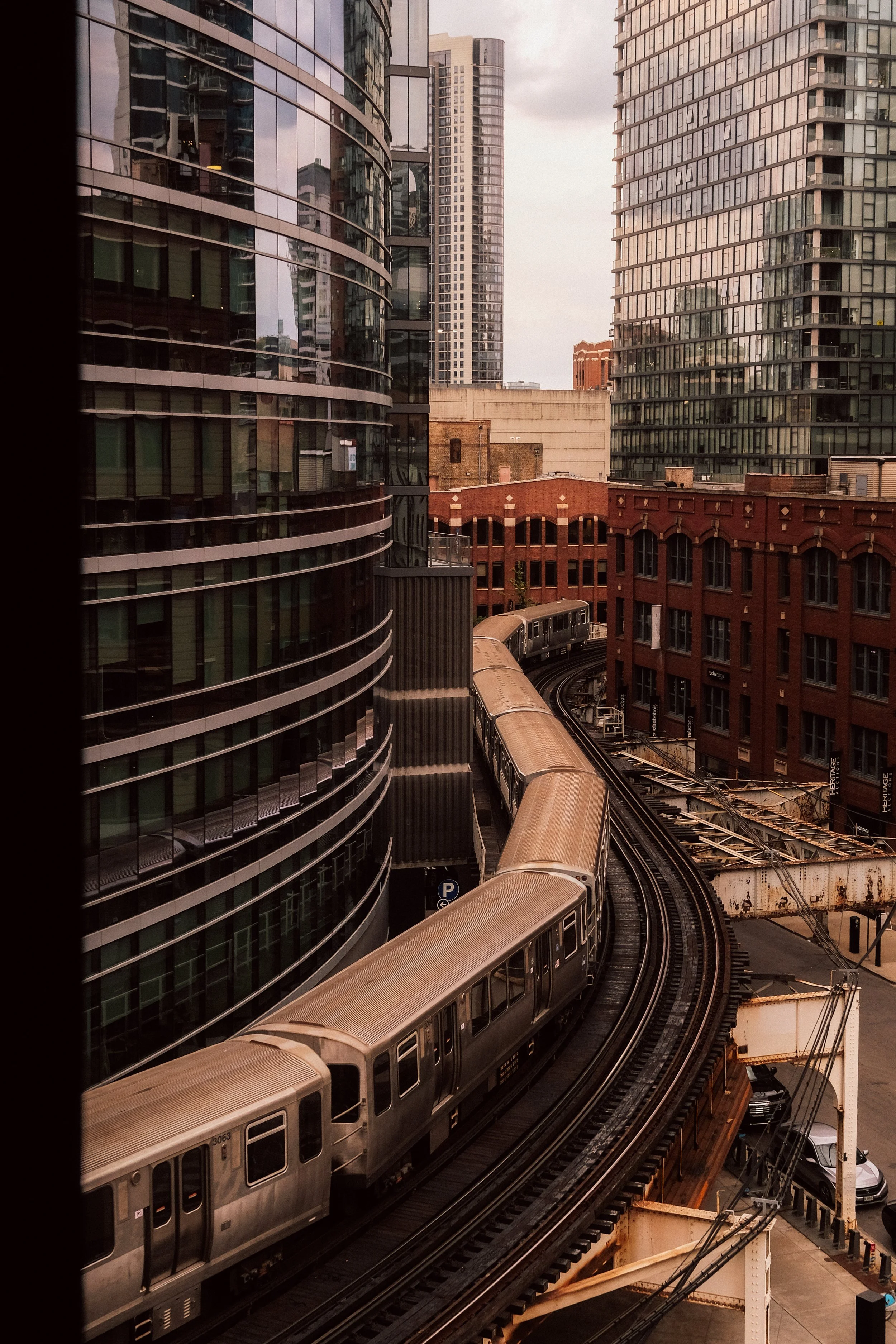 A city scene with curved glass skyscrapers, an elevated train on tracks, and old brick buildings in the background.