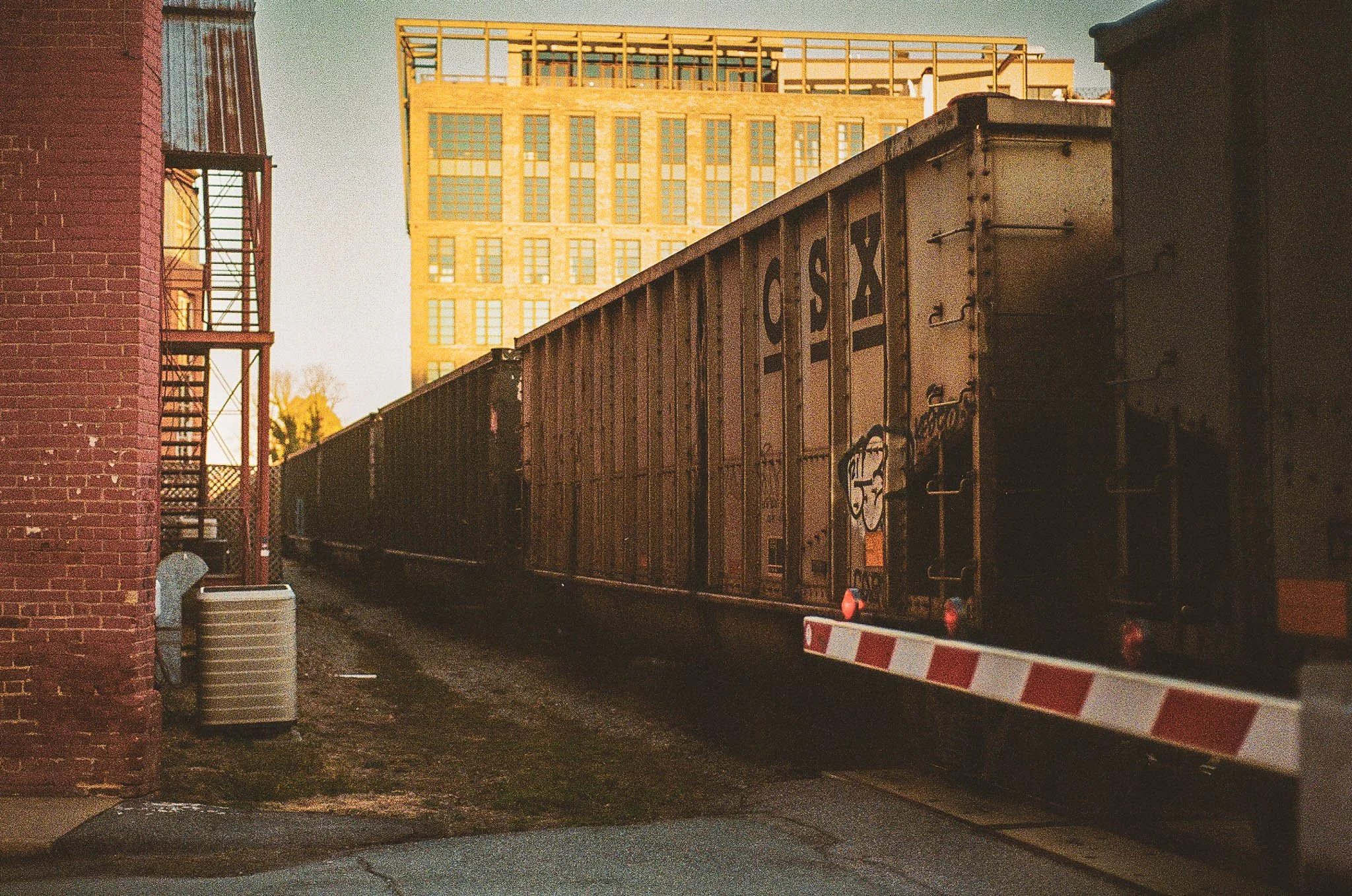 A freight train passing by a brick building with a fire escape on the side, with a tall modern building in the background, during sunset.