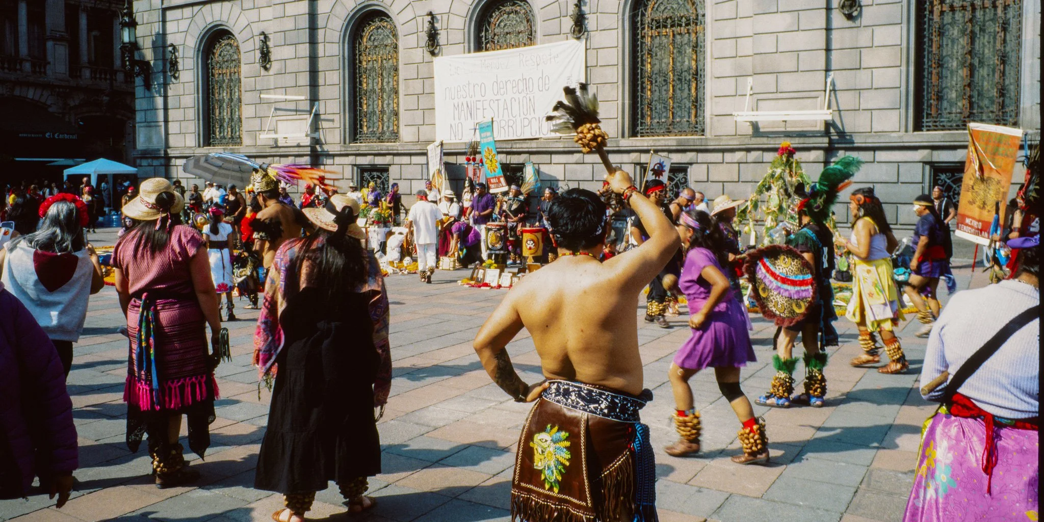 People participating in a traditional cultural dance outdoors, with some dressed in colorful costumes, and a building with barred windows in the background.