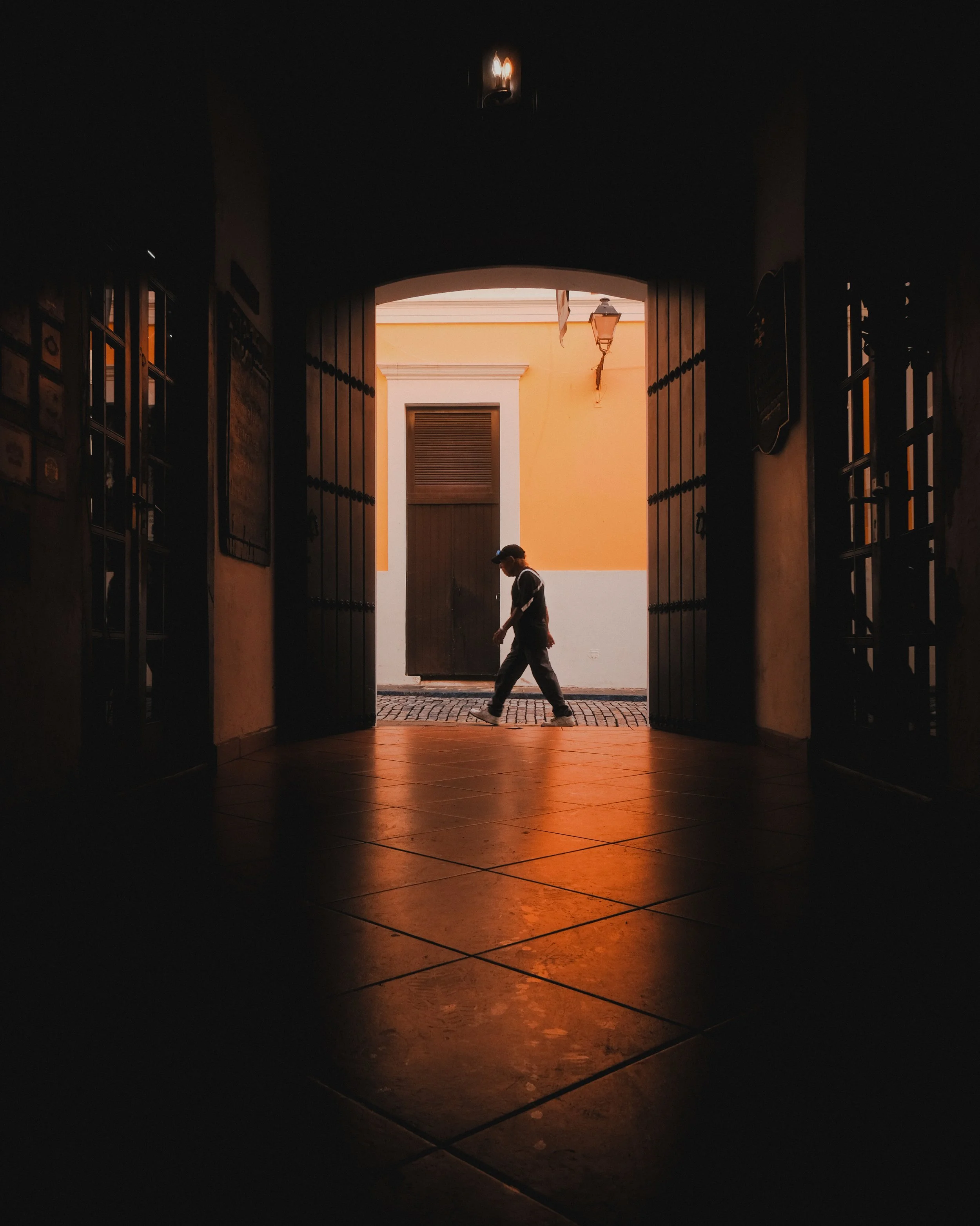 A person walking outside through an open doorway with a yellow wall and orange sky in the background, viewed from inside a dark room.