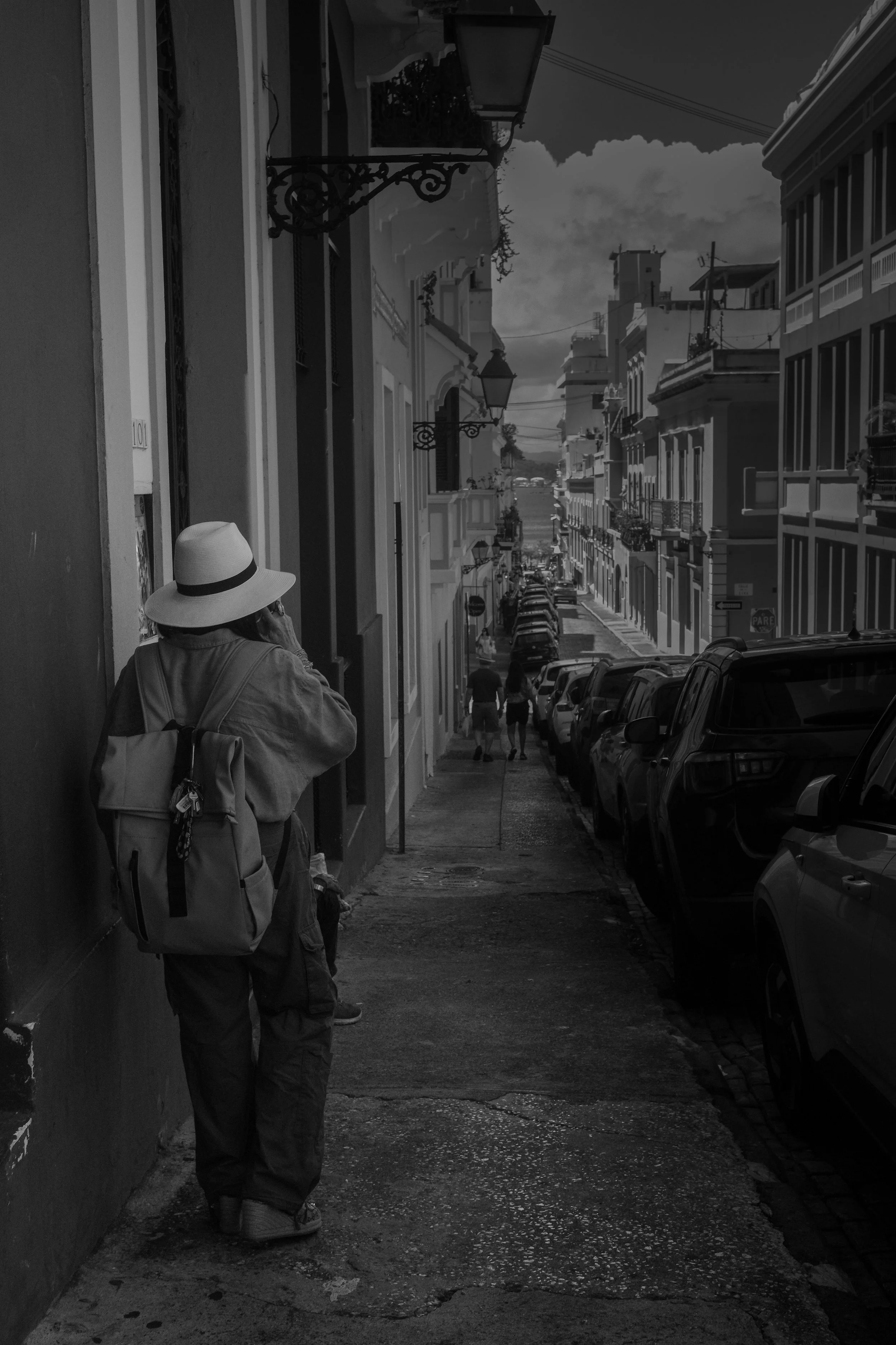 A person with a backpack and hat standing on a city sidewalk, talking on the phone. The street is lined with parked cars and historic buildings with ornate street lamps.