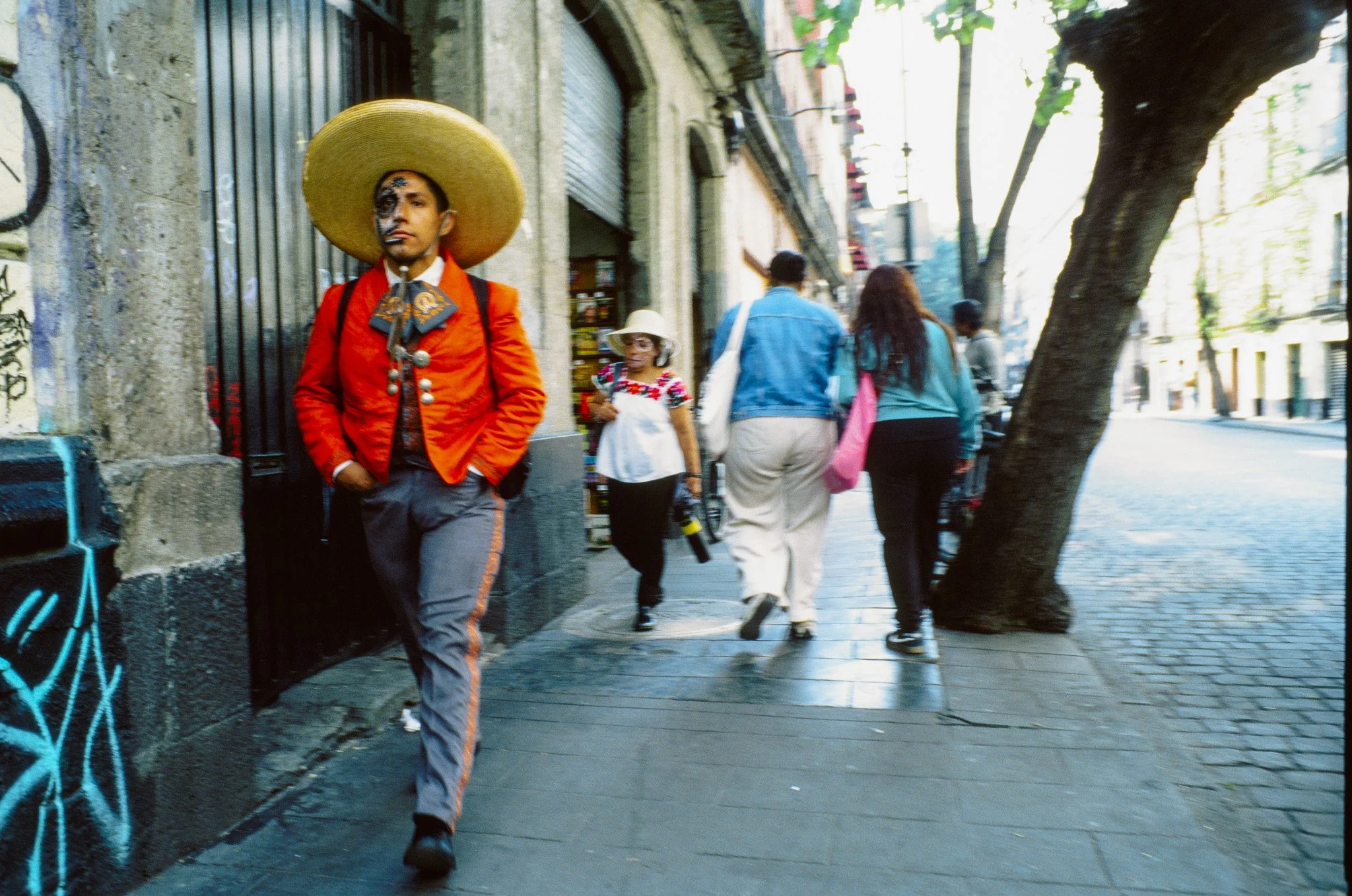 A man dressed as a Mexican mariachi, walking on a city sidewalk, wearing a large sombrero and traditional colorful costume, with face paint.