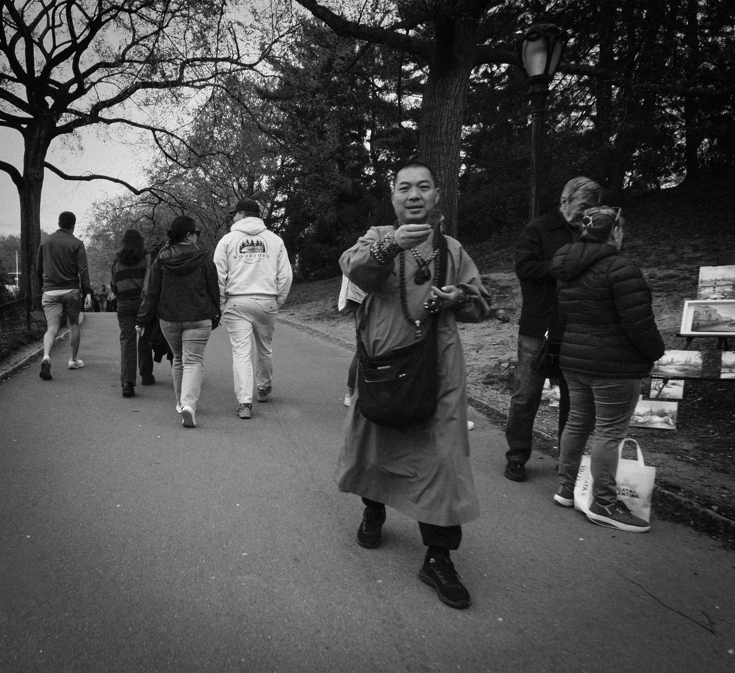 A group of people walking along a park pathway with trees on either side, some viewing artwork displayed on stands, and one man in the foreground gesturing towards the camera.