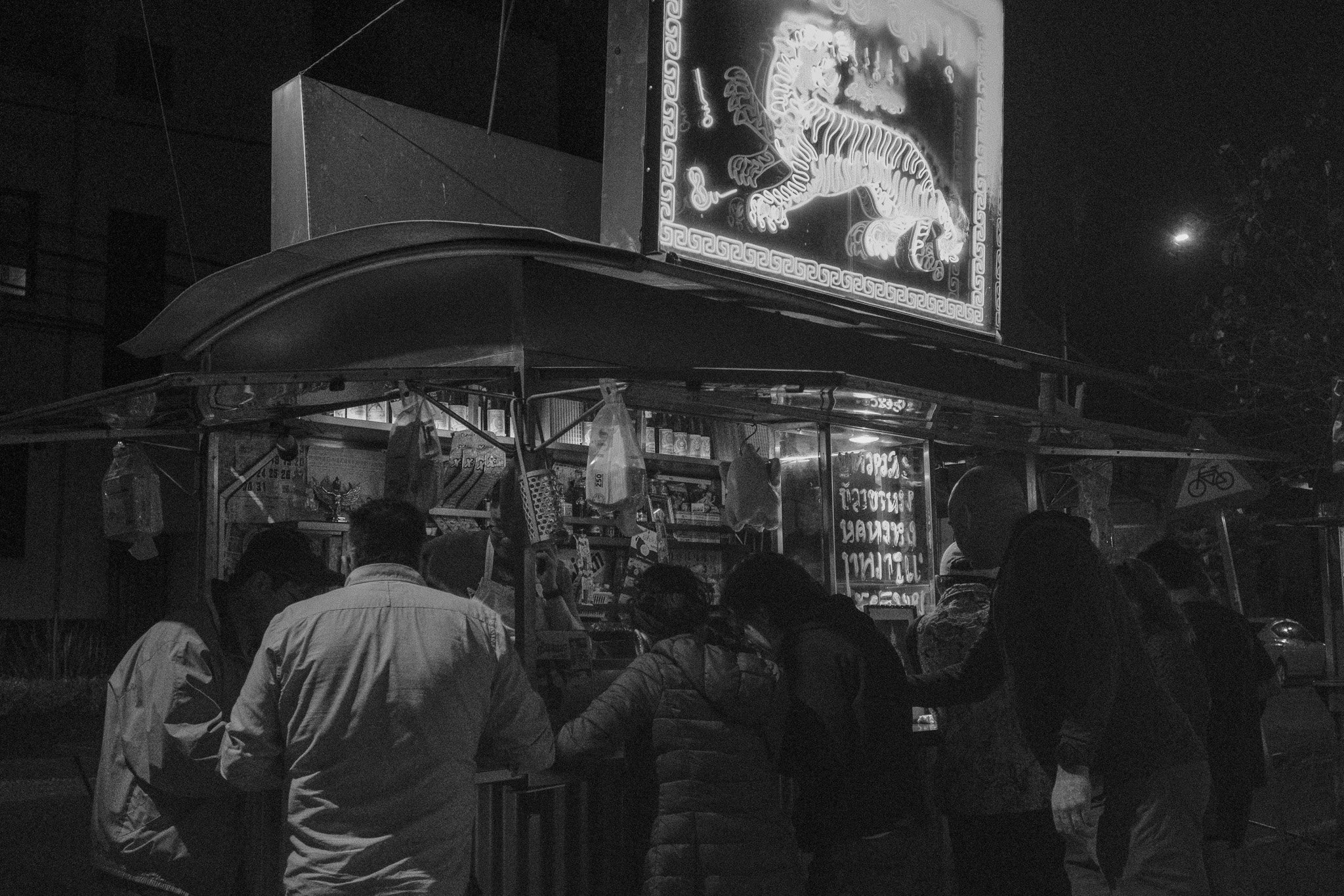 Night scene of a street food cart with people gathered around, illuminated by a neon sign featuring a tiger at the top.