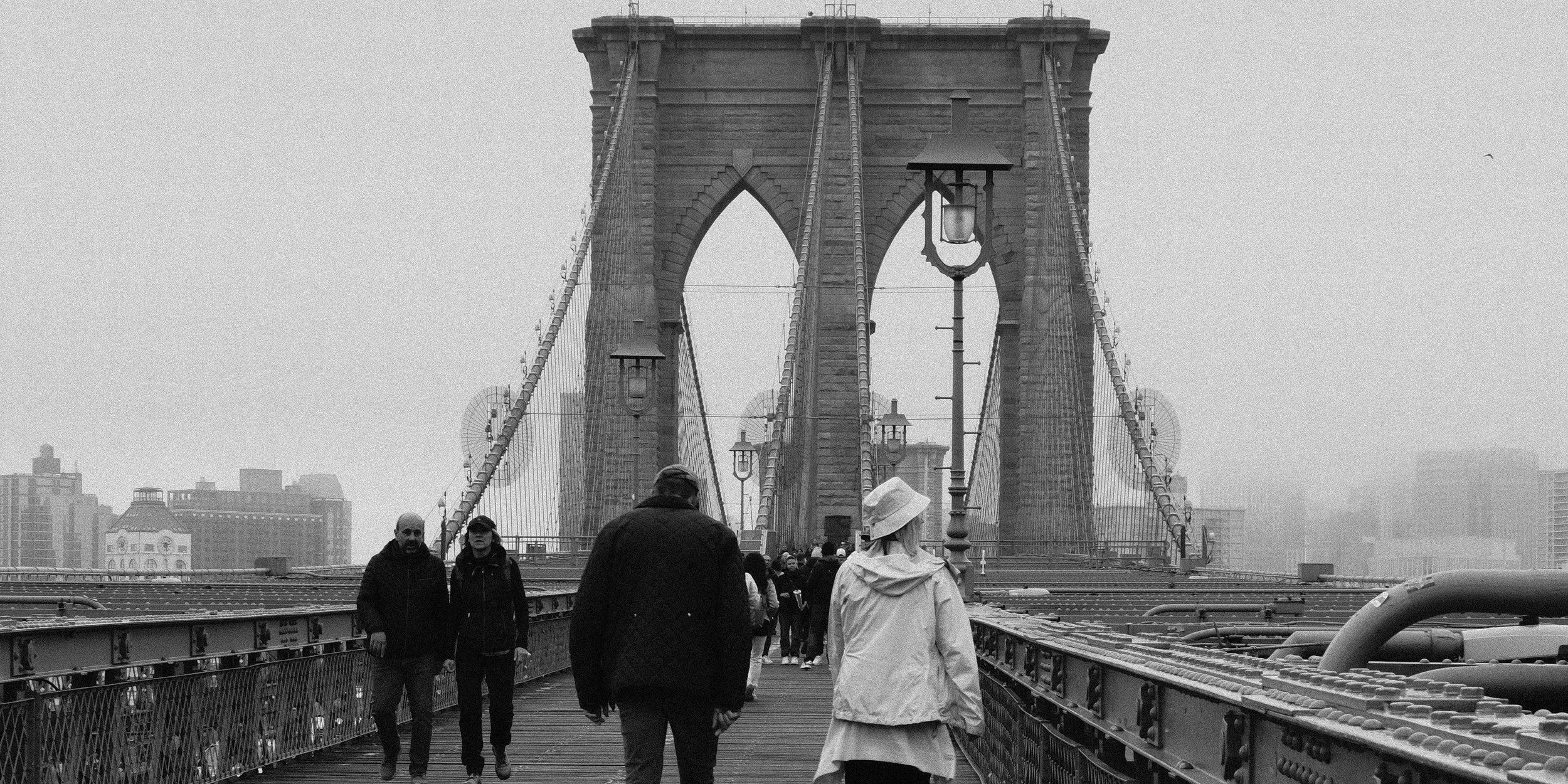 People walking on the Brooklyn Bridge in New York City, with city buildings in the background, captured in black and white.