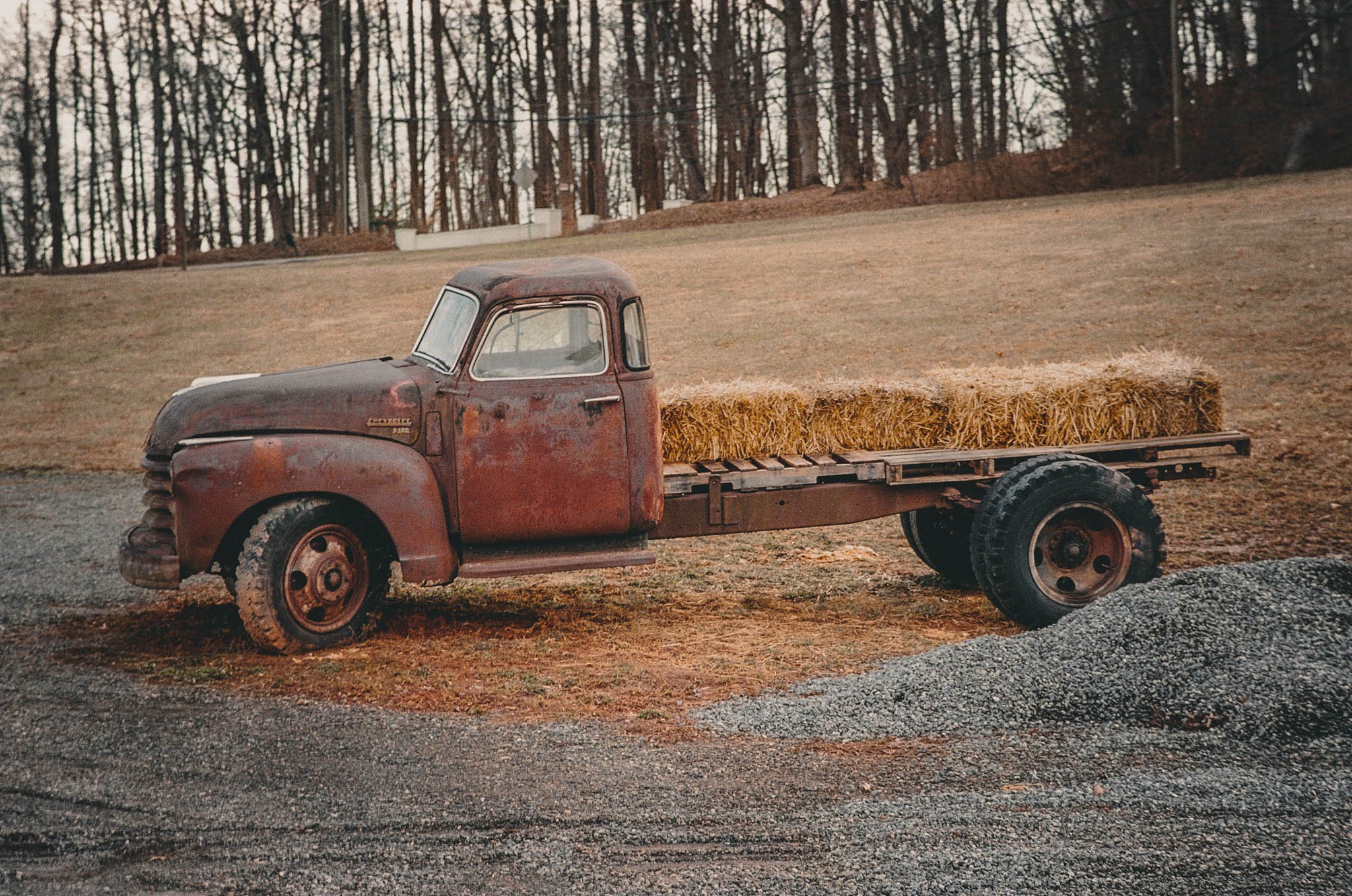 An old rusty vintage truck with a flatbed loaded with hay bales, parked on dirt and gravel ground with a background of leafless trees on a cloudy day.